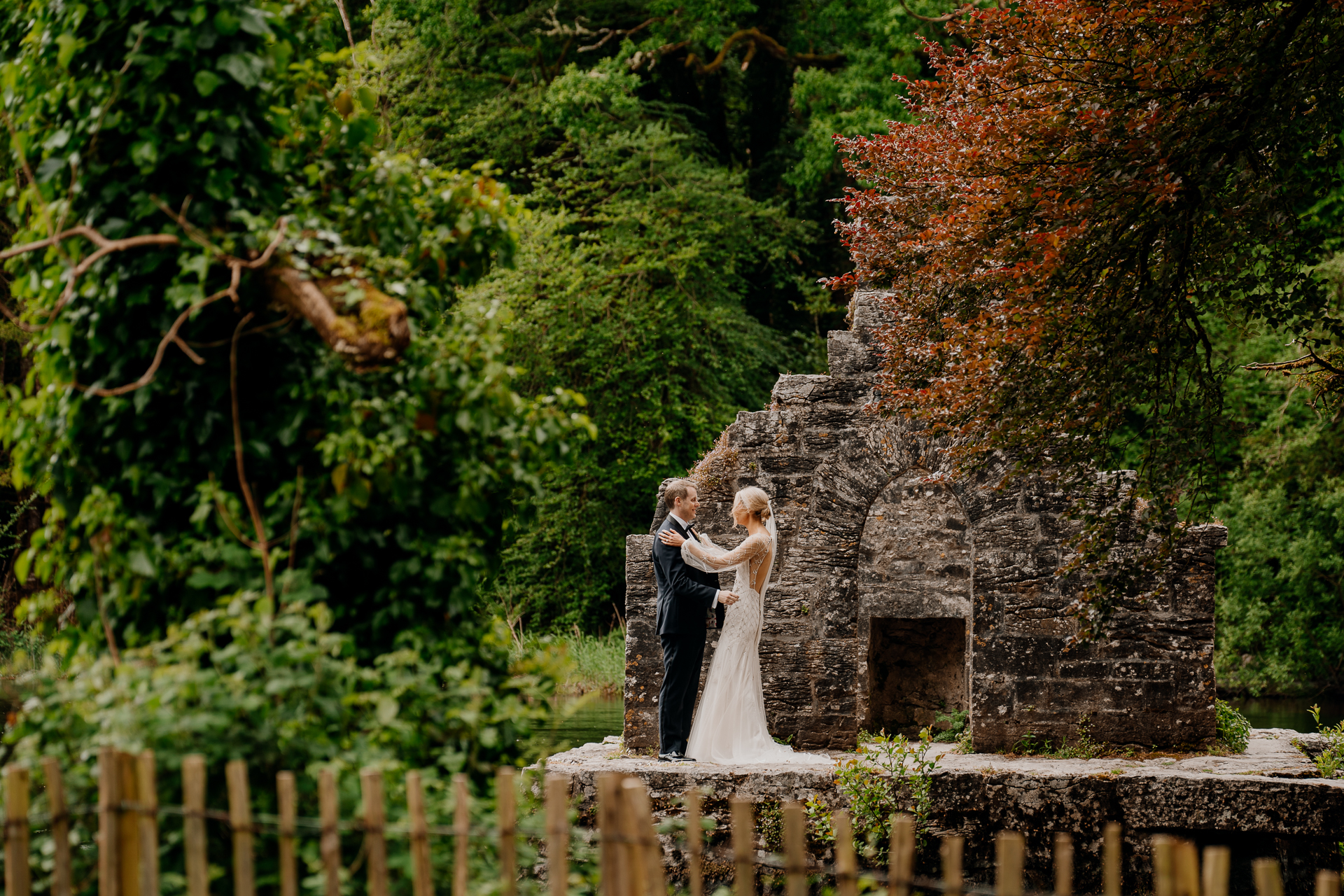 A couple kissing in front of a stone building with trees and a fence