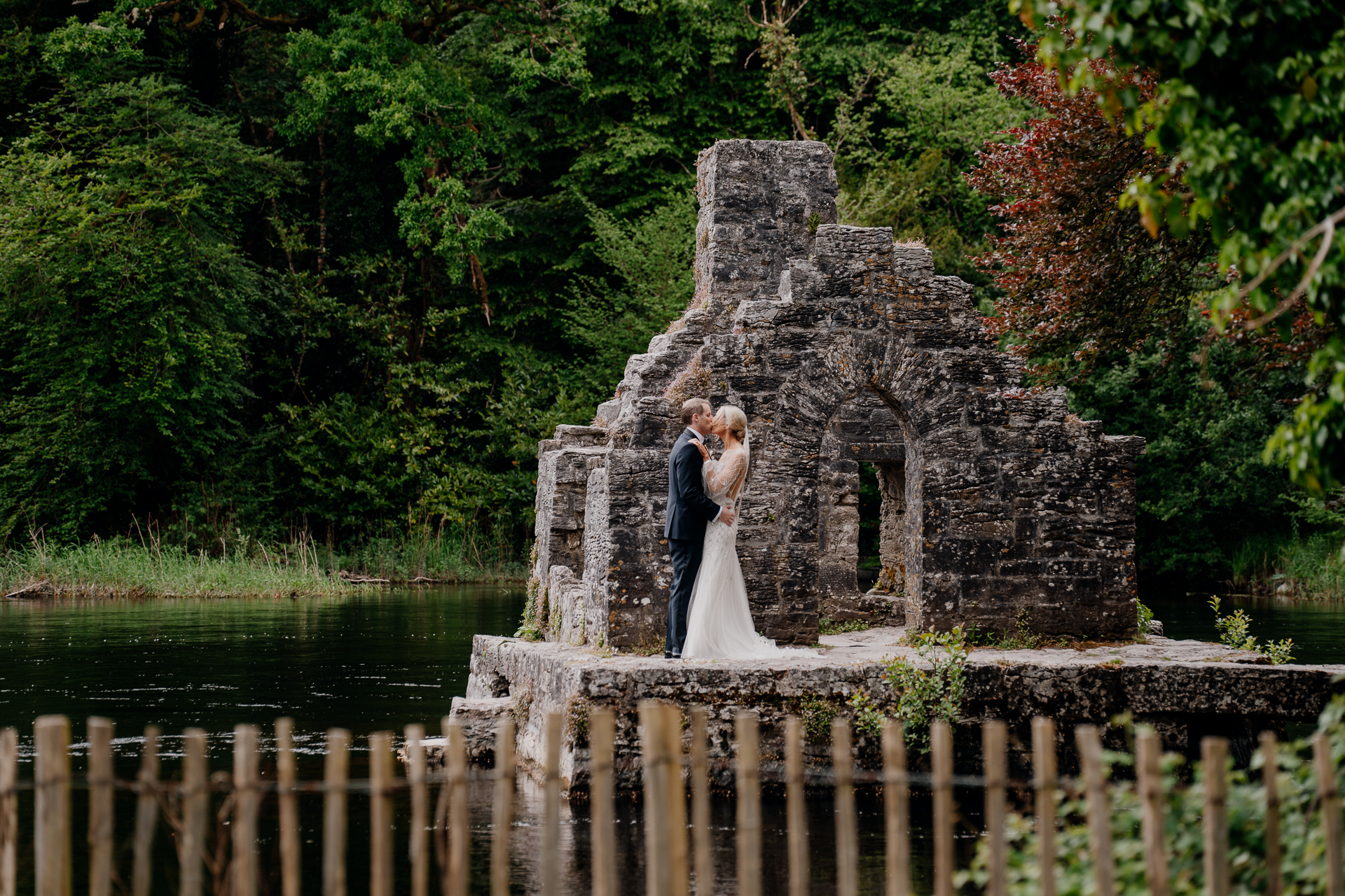 A couple kissing in front of a stone structure with trees in the background