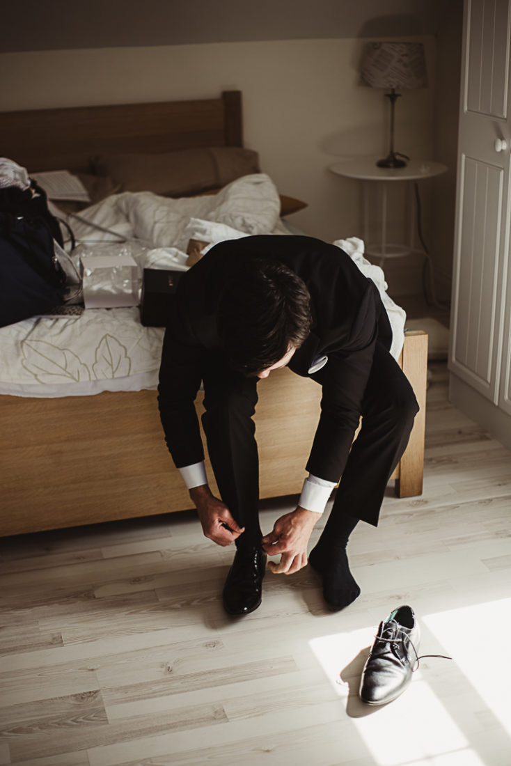 A groom sitting on a bed wearing shoes before the wedding