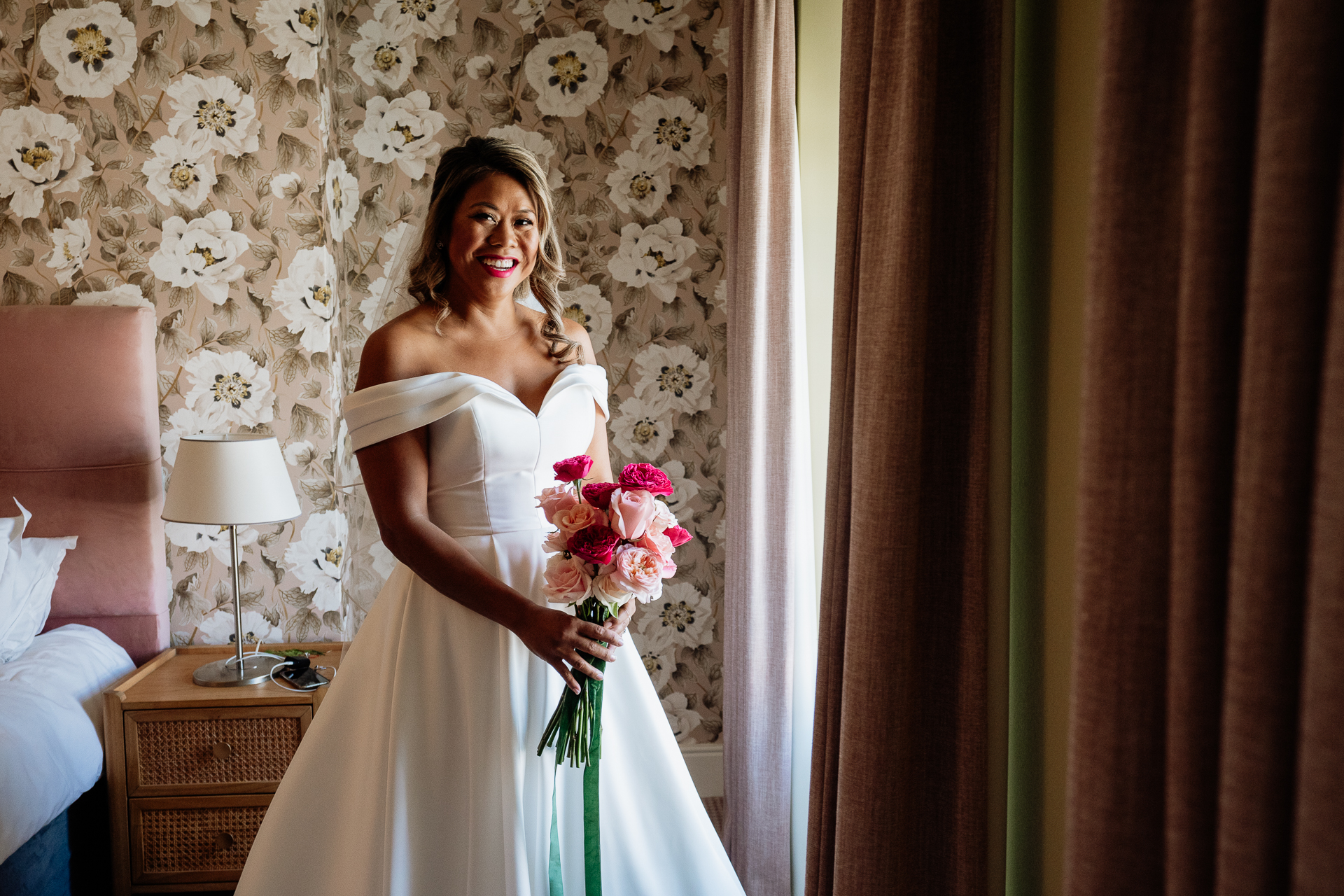 A person in a white dress holding flowers in a room with a wallpaper