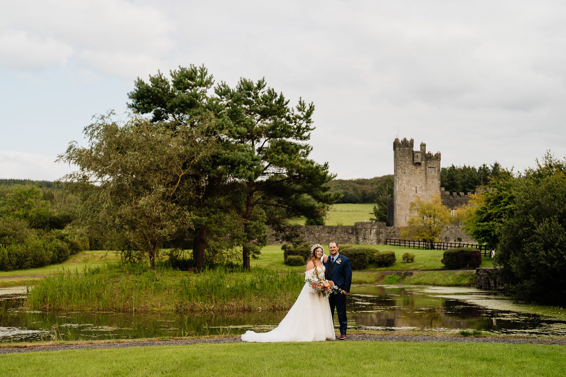 A man and woman in wedding attire standing in a grassy field with a castle in the background