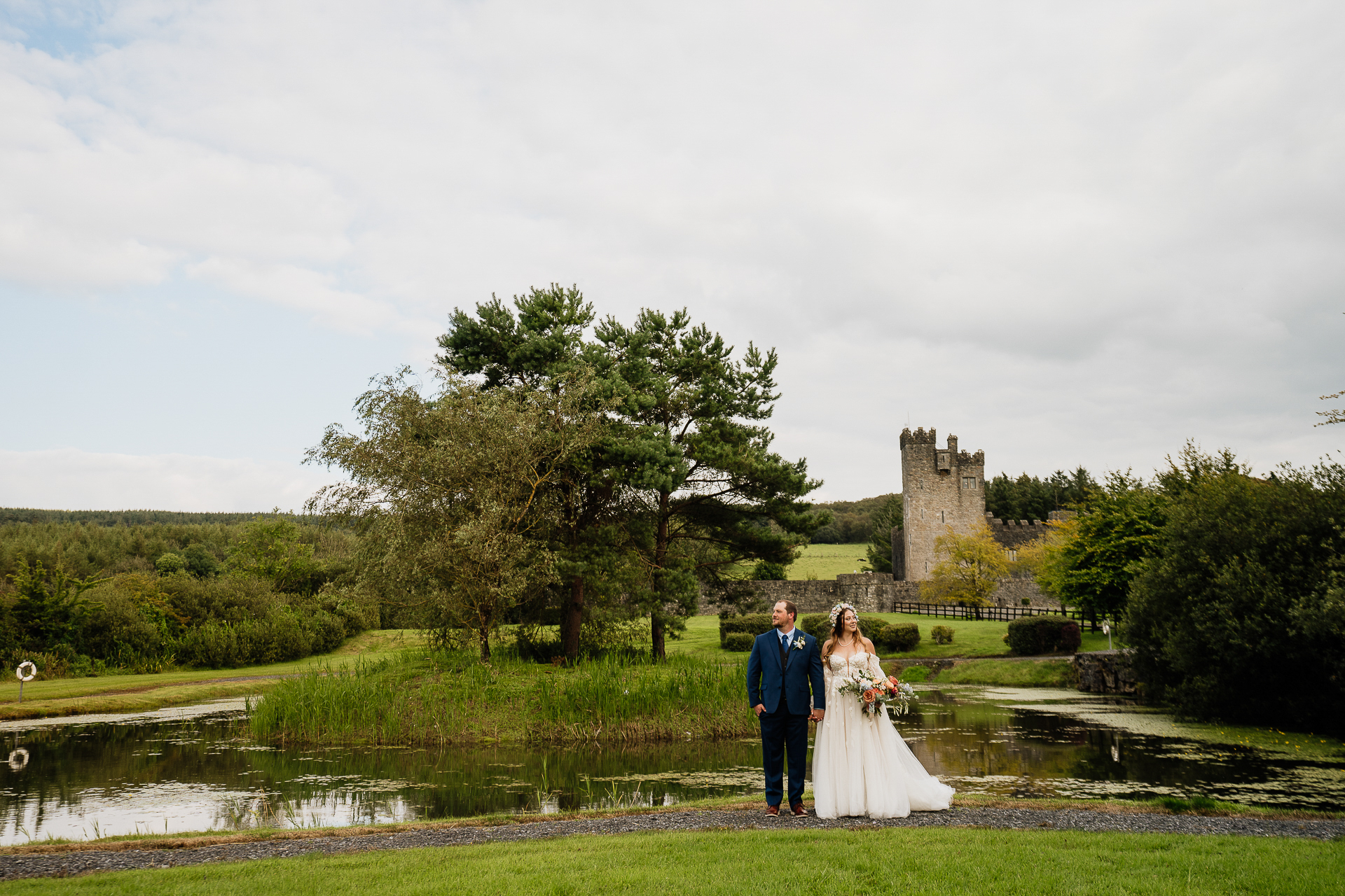 A man and woman in wedding attire standing in a grassy field with a castle in the background