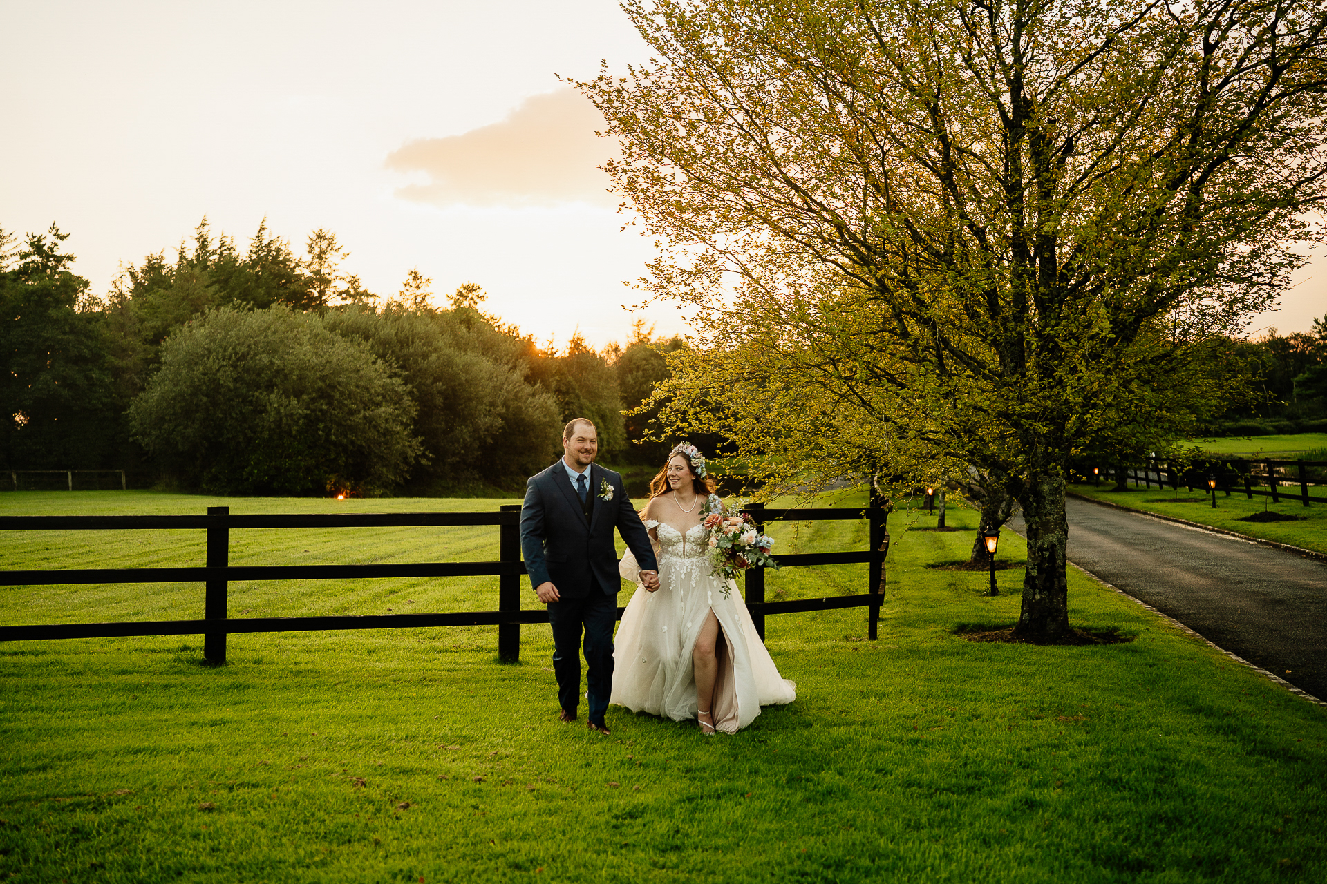 A man and woman in wedding attire standing in a grass field with trees and a fence and a