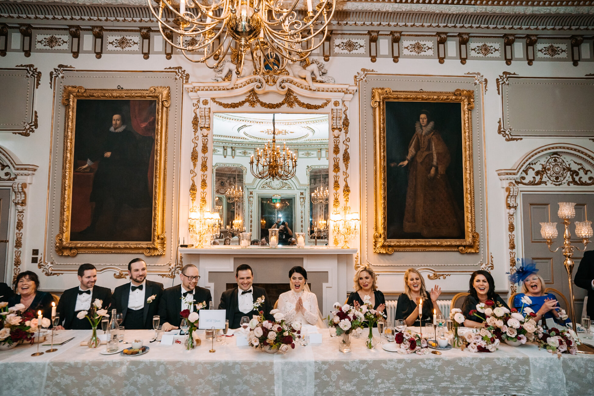 A group of people sitting at a table with flowers in front of a painting