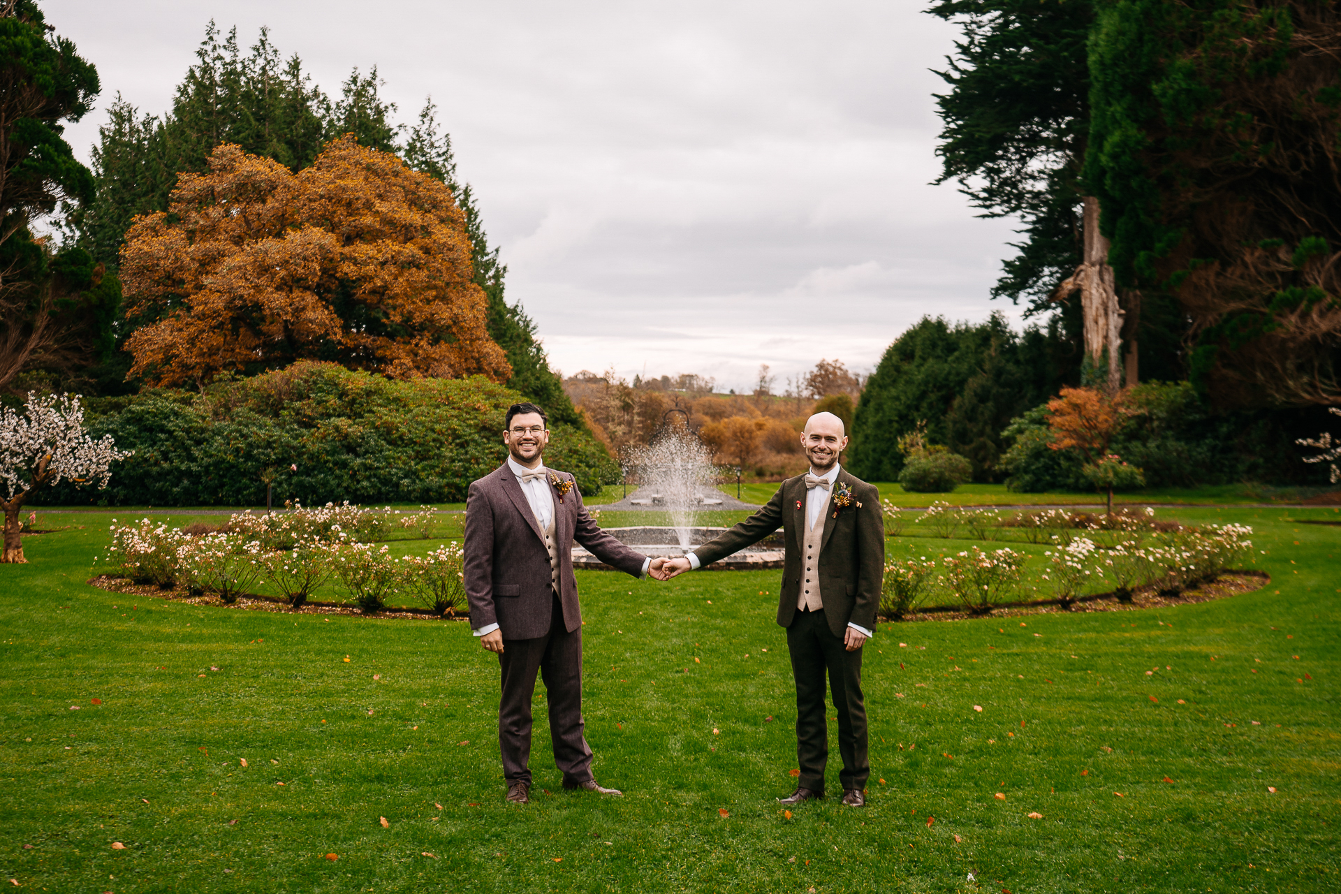 Two men in suits standing in a grass field with a fountain in the background