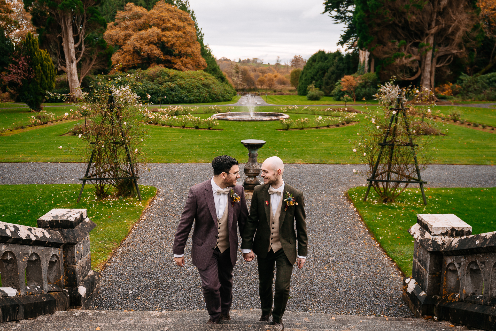 A couple of men in suits walking down a path with a fountain