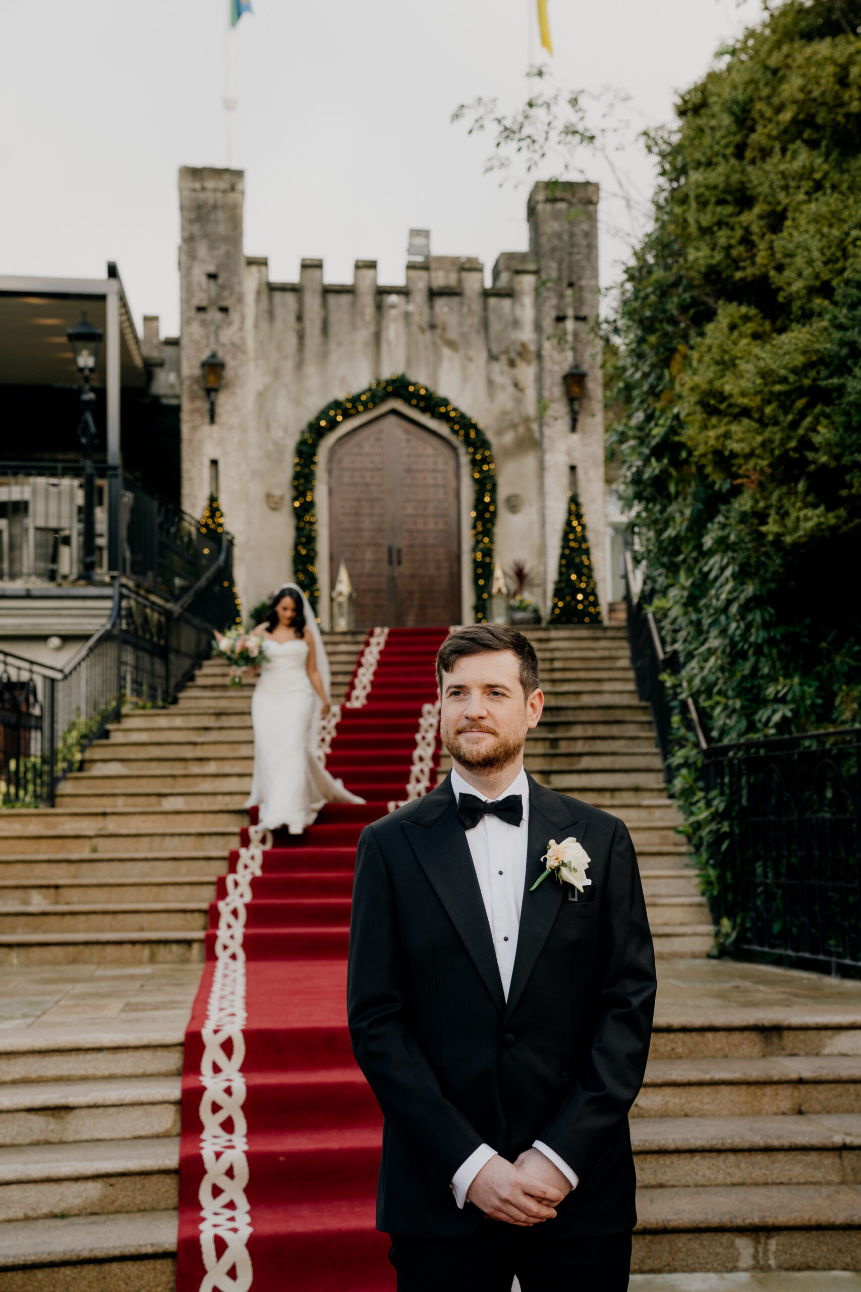 A man in a tuxedo standing on stairs with a woman in a dress in the background