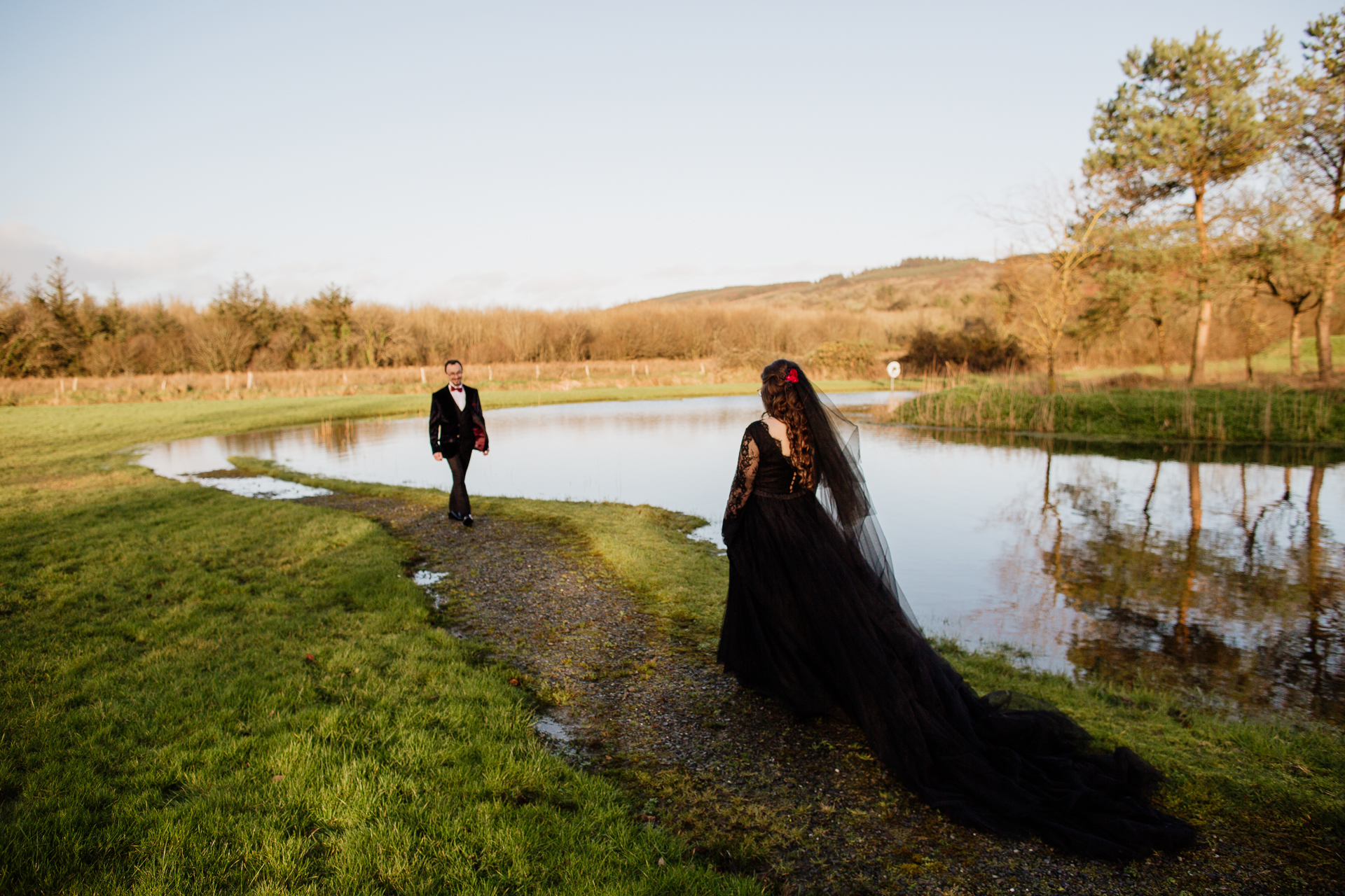 A man and woman in a dress standing on a dirt path by a pond with trees and grass