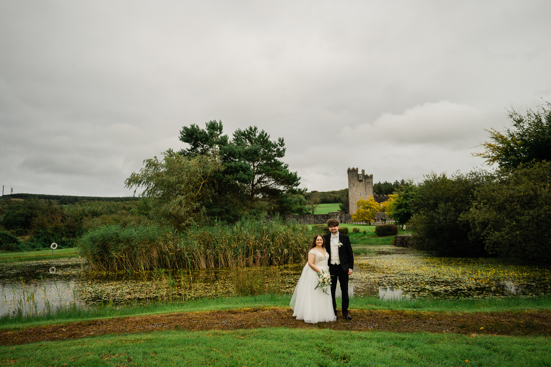 A man and woman in wedding attire standing in a field with a pond and trees and a castle