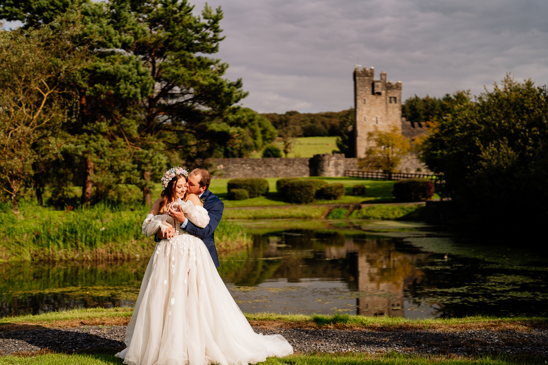 A man and woman in wedding attire kissing by a pond with a castle in the background