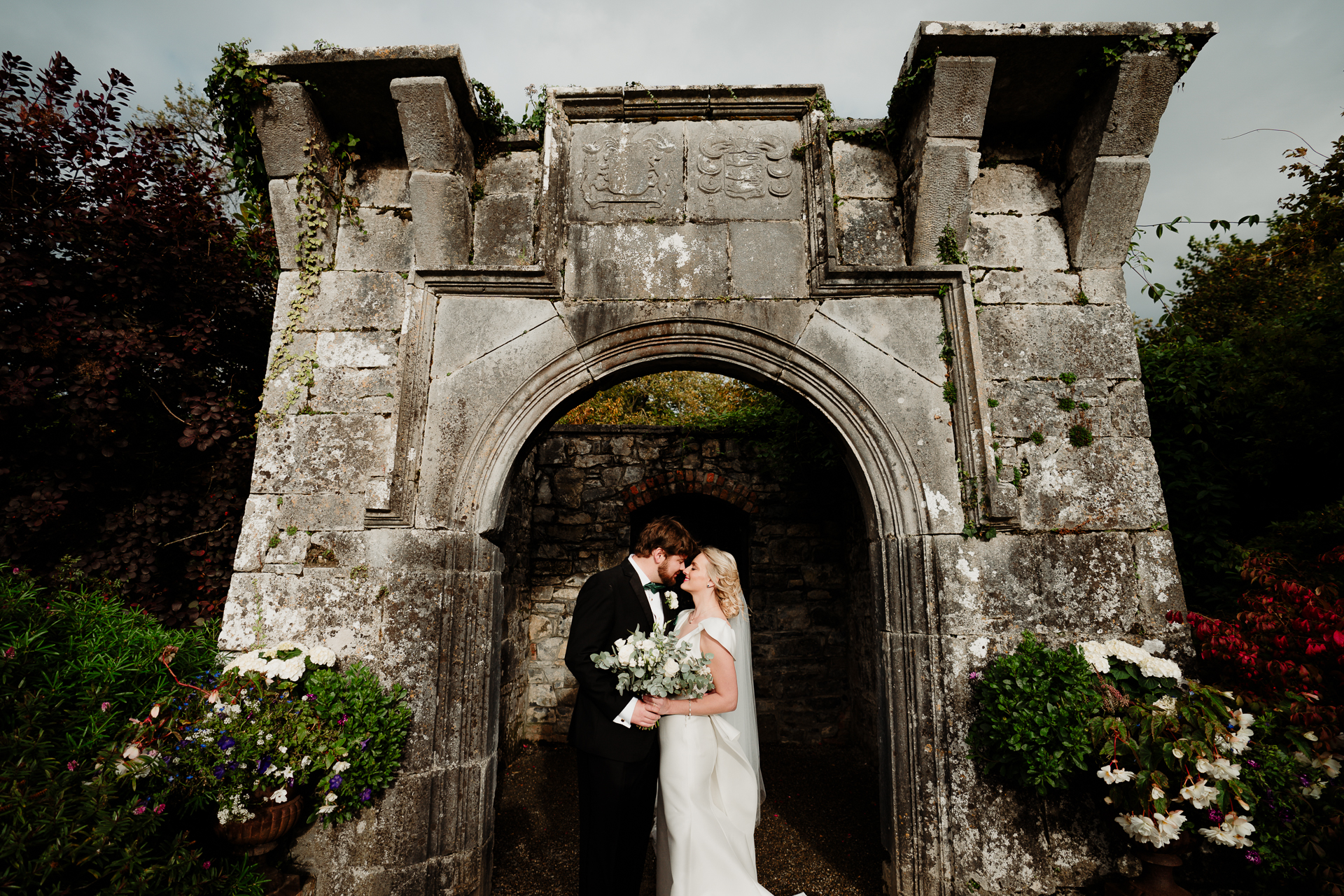 A man and woman in wedding attire kissing in front of a stone archway