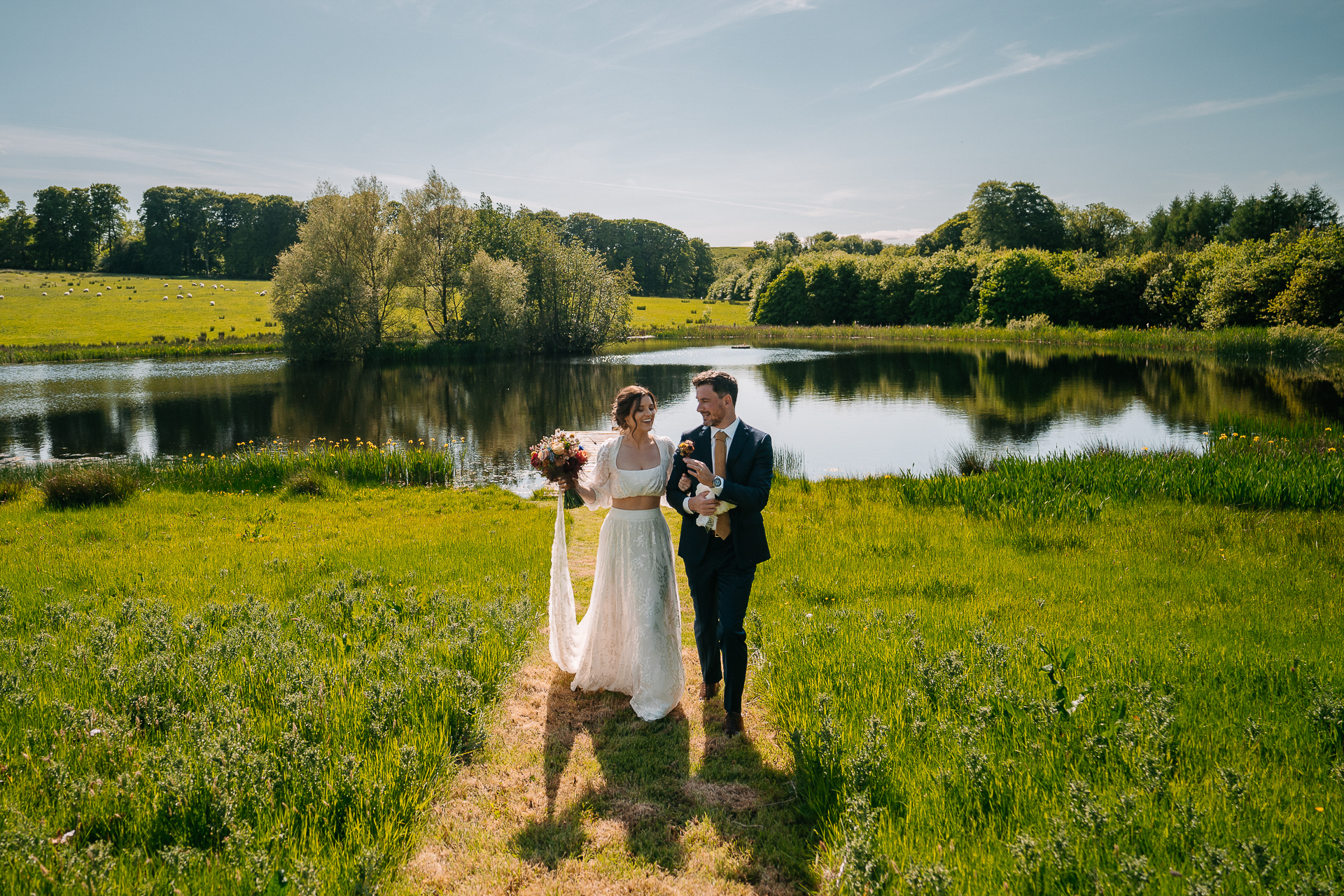 A man and woman in wedding attire standing on a dirt path by a pond with trees and grass