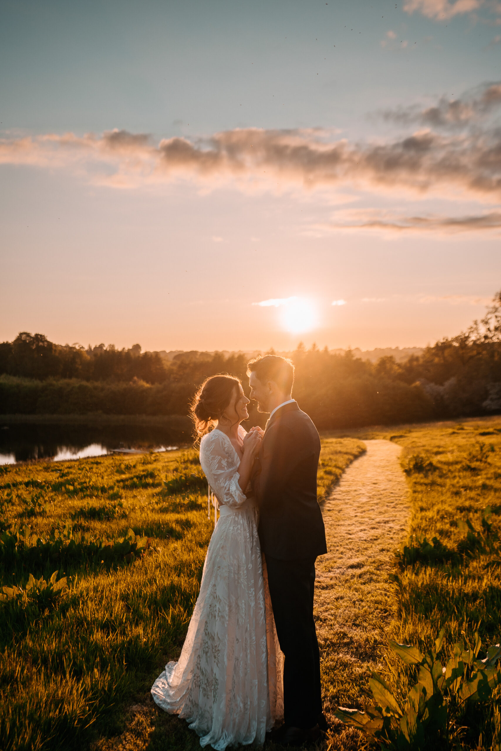 A man and woman kissing on a path with grass and trees and a sunset