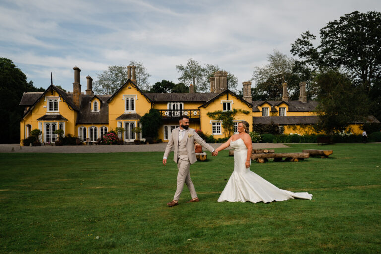 A man and woman in wedding attire walking on a lawn in front of a large house