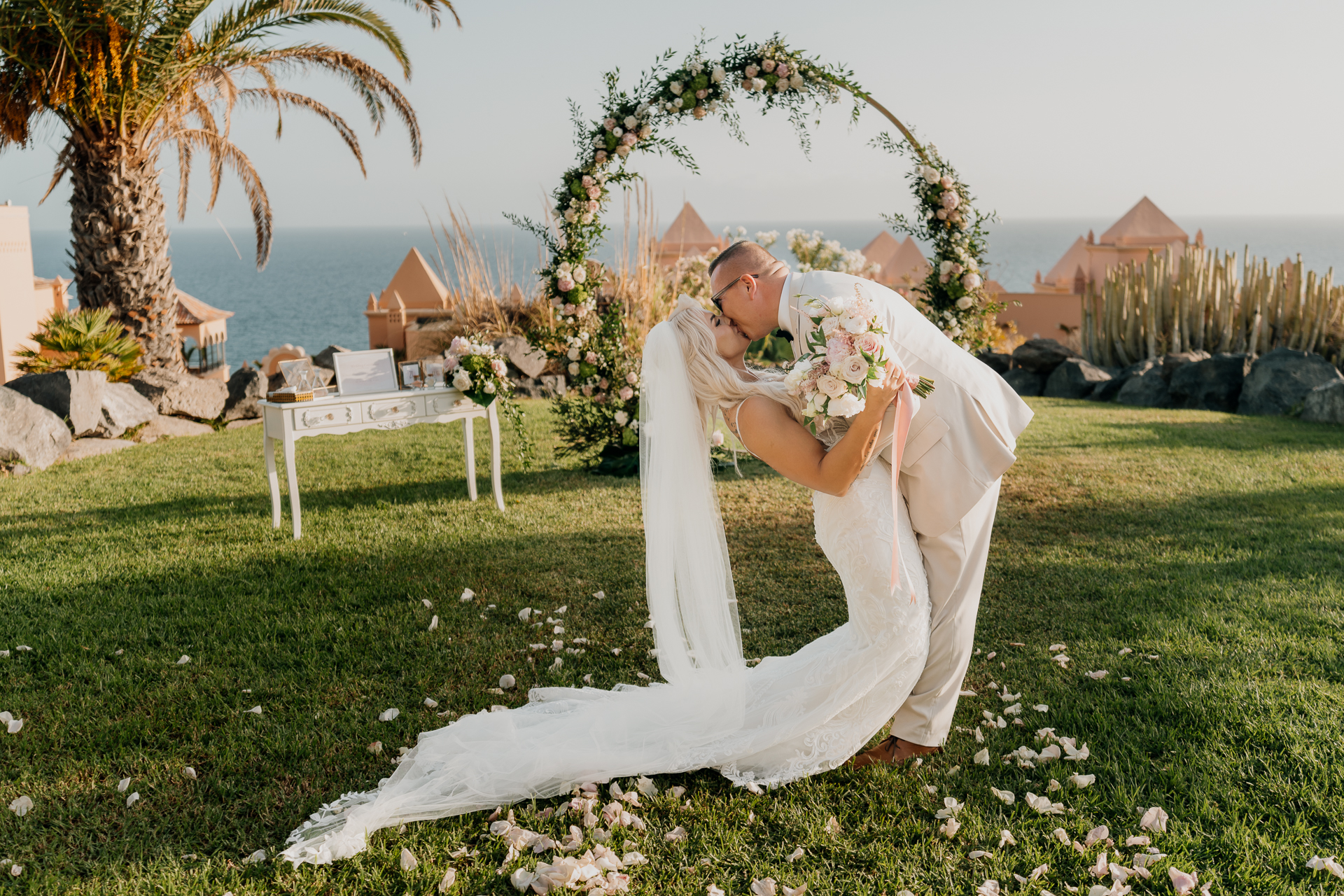 A person in a wedding dress sitting on a lawn with a table and chairs and a beach and