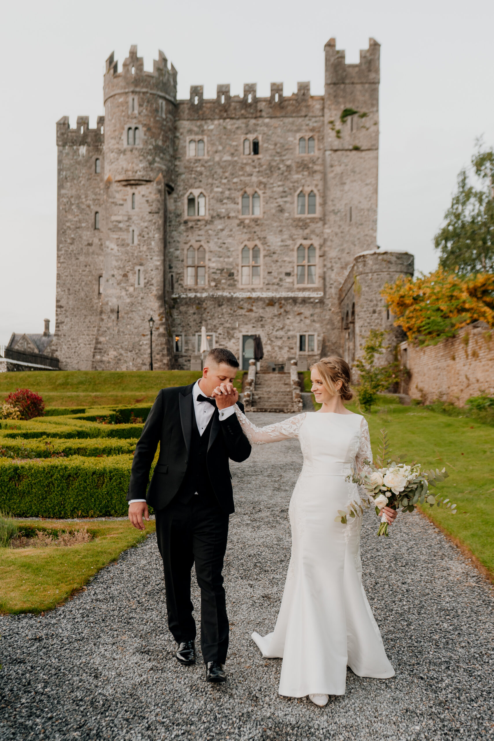 A man and woman in wedding attire walking down a path in front of a castle