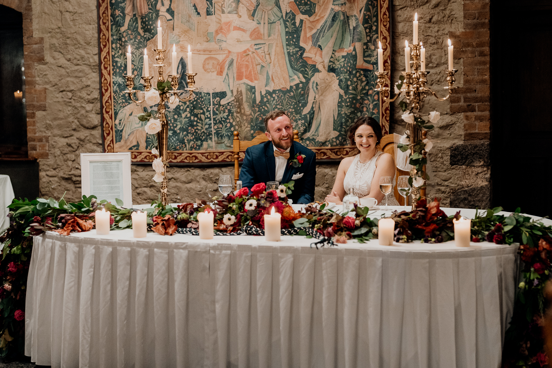 A bride and groom sitting at a table with flowers and candles