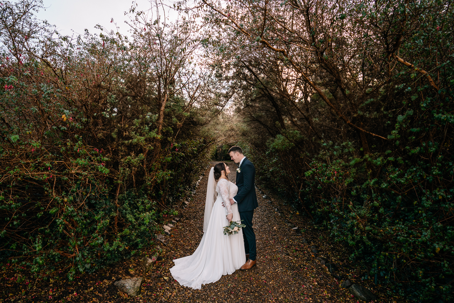 A man and woman in wedding attire walking down a path with trees