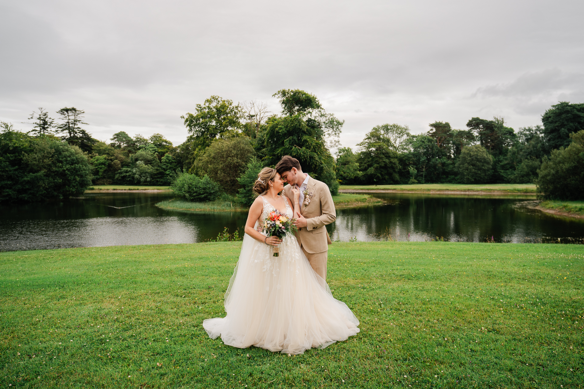 A man and woman kissing in a grass field with a pond and trees