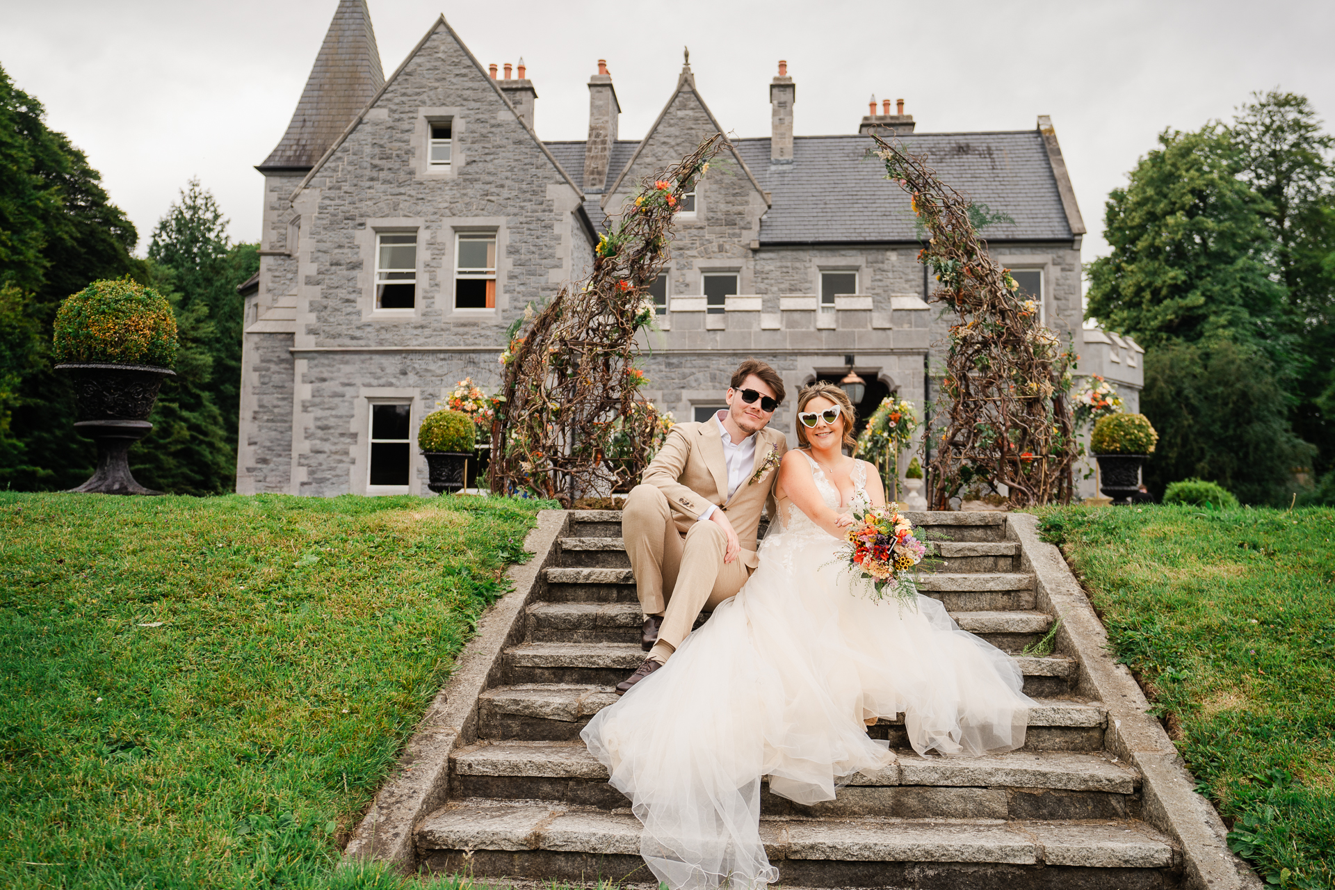 A man and woman in wedding attire sitting on a white bench in front of a house