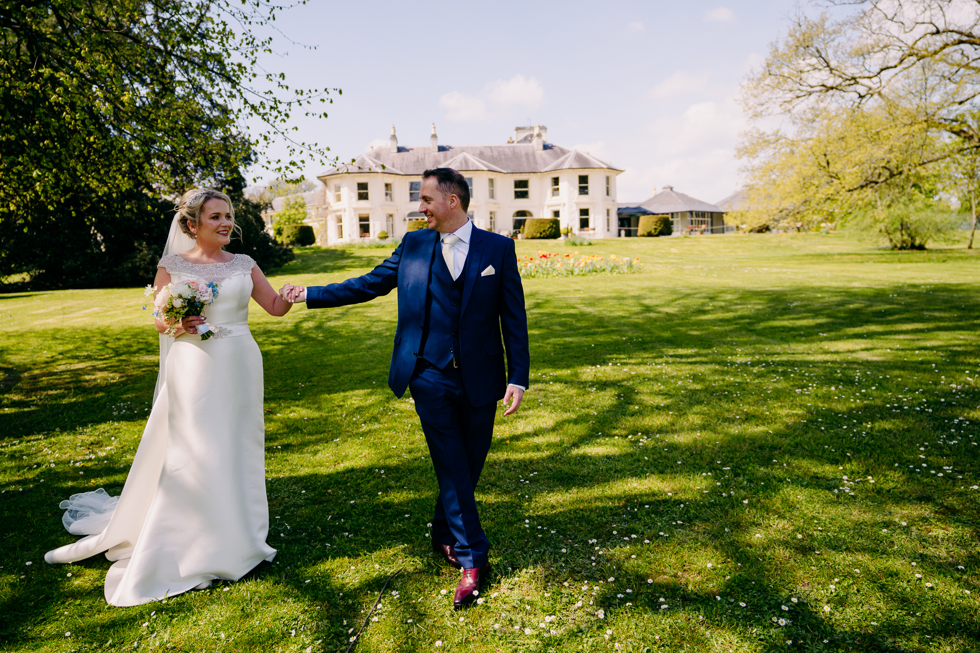 A man and woman in wedding attire walking on grass with a building in the background