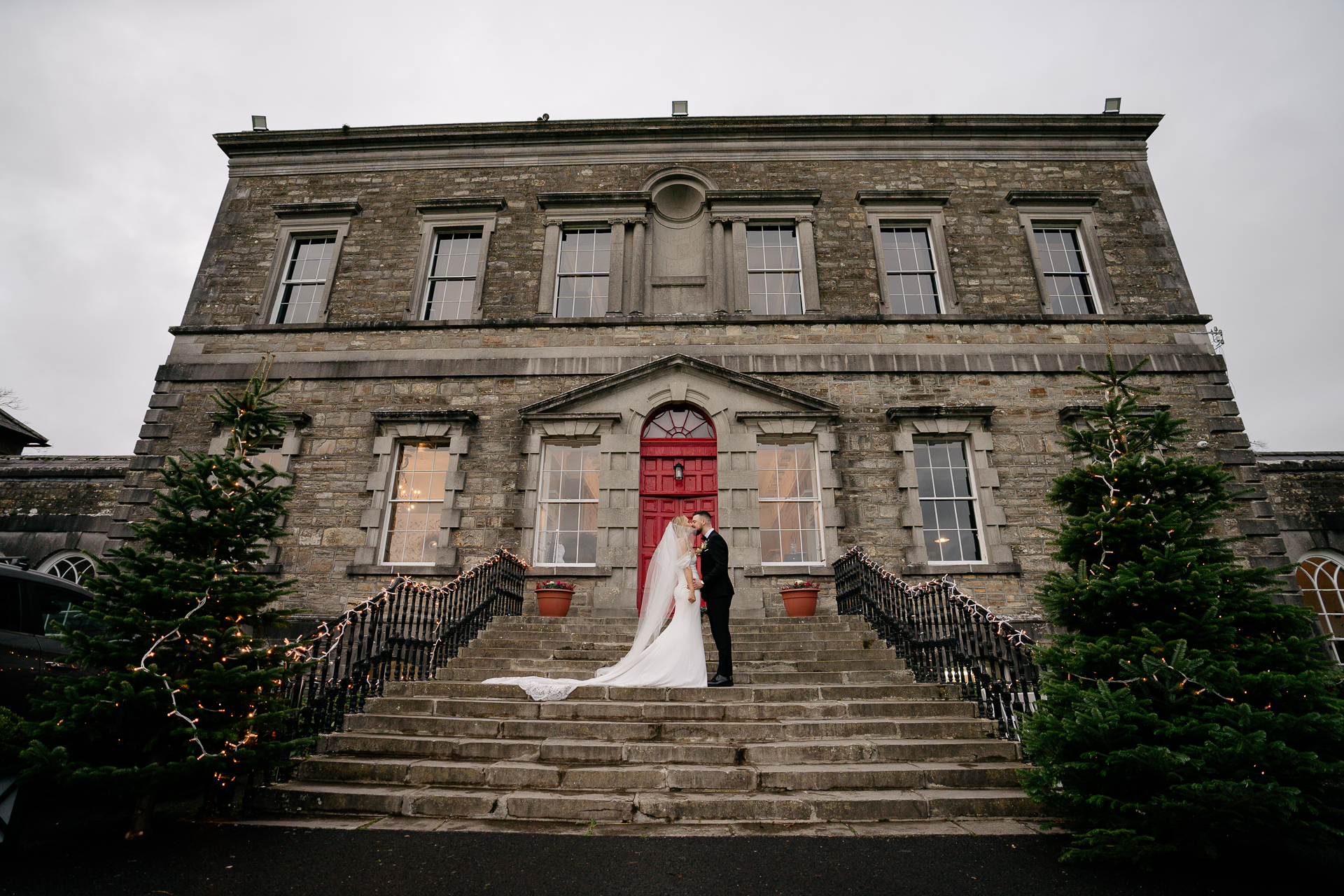 A bride and groom standing on the steps of a large house