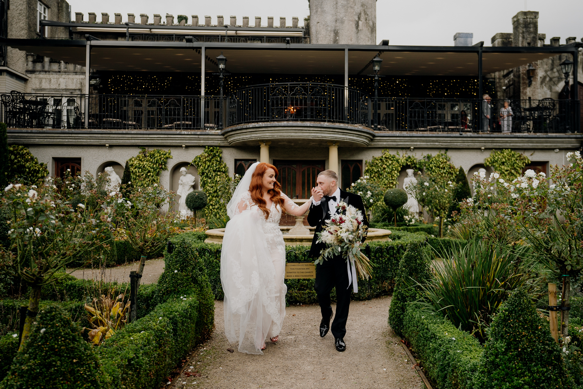 A man and woman in wedding attire walking down a path in front of a building with many plants