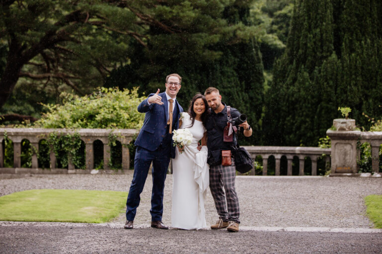 Wojciech Koza, a wedding photographer, standing with a bride and groom holding a camera in front of a scenic background at Belleek Castle