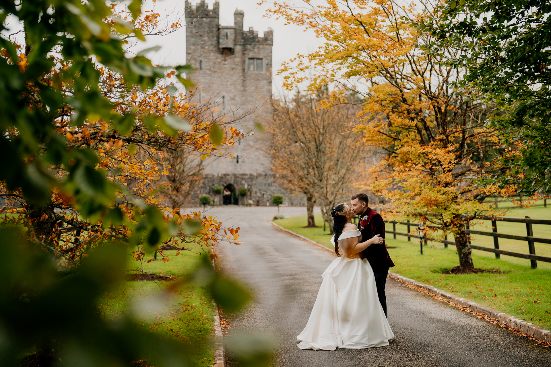 A man and woman kissing on a path with trees and a building in the background