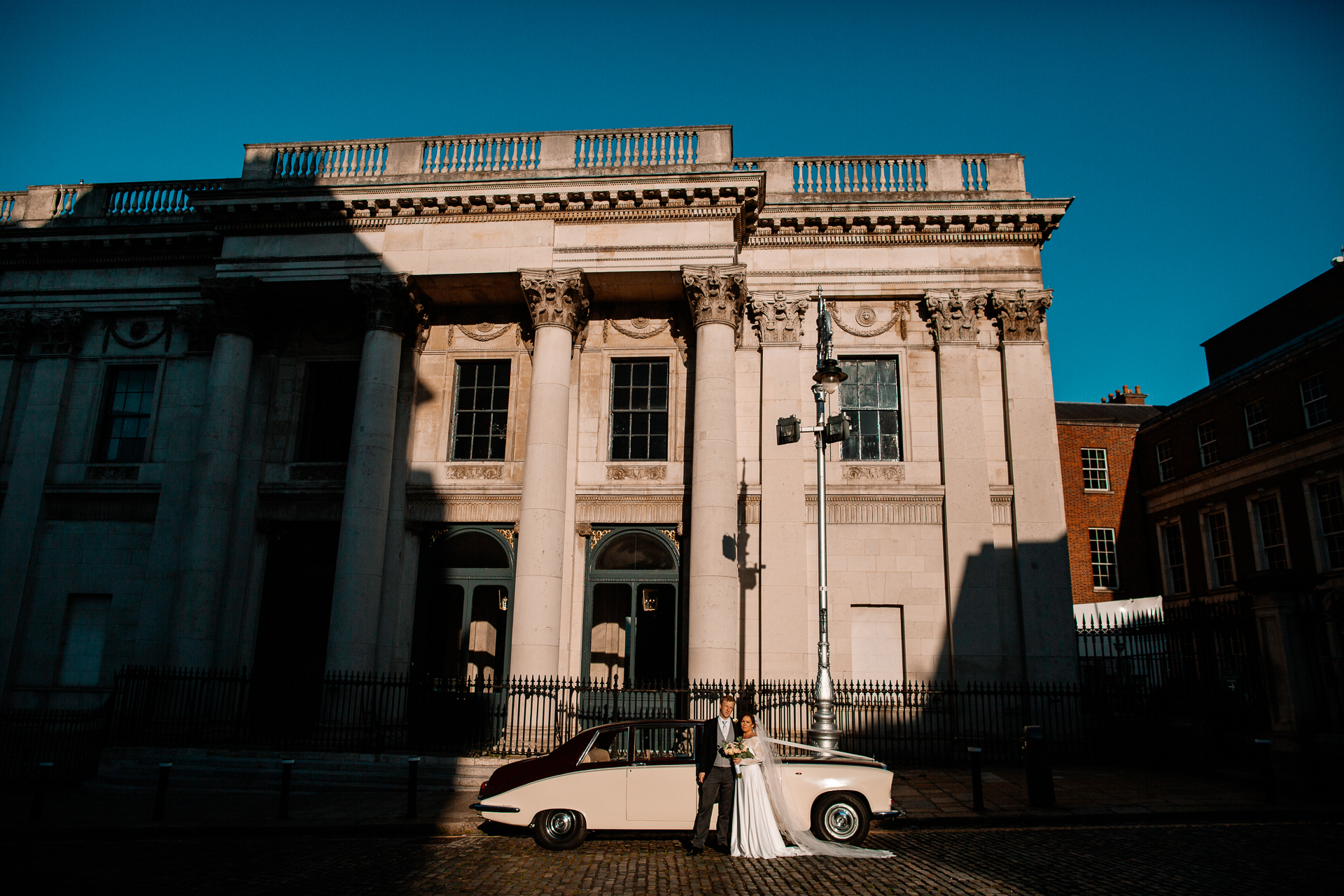 A bride and groom standing in front of a white car in front of a building