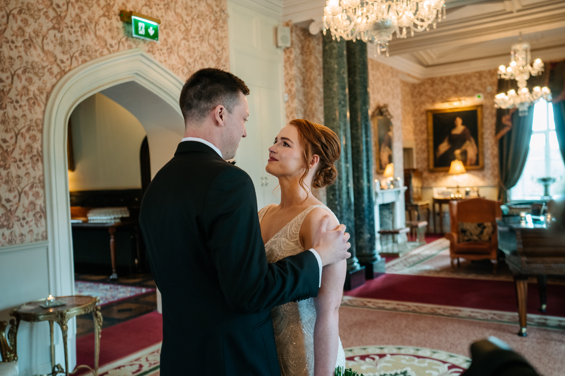 A man and woman kissing in a room with chandeliers and a fireplace