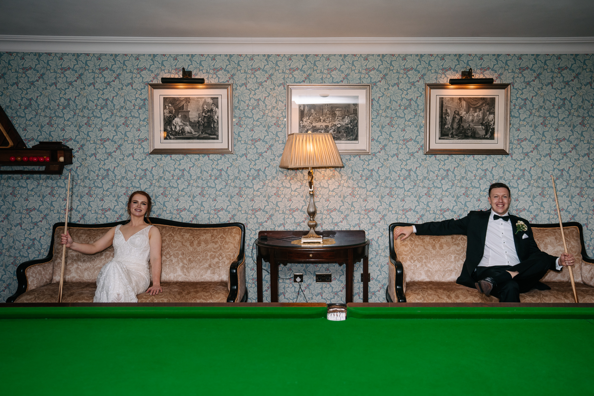 A man and woman sitting on a couch in a room with a pool table and a lamp