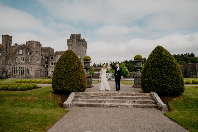 A bride and groom walking down a stone staircase in front of Ashford Castle, Ireland, surrounded by lush gardens.