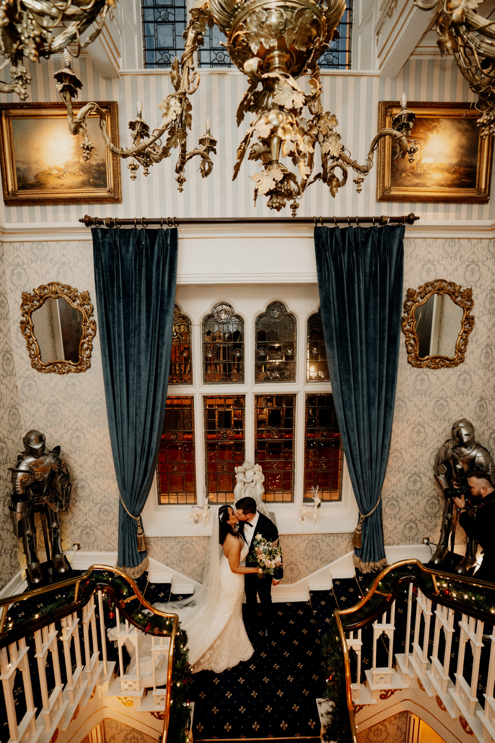 Newlyweds standing on the main staircase inside Cabra Castle under golden chandeliers.