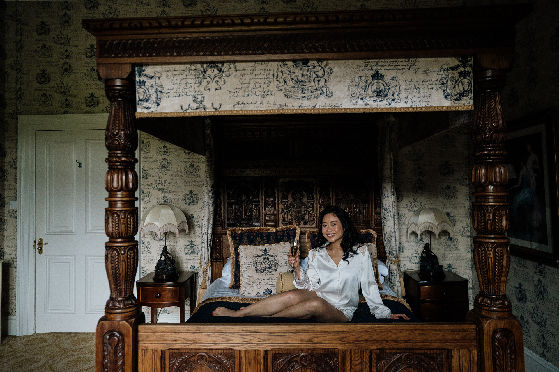 The bride relaxing on a four-poster bed at Belleek Castle in a white robe, surrounded by vintage wallpaper and castle decor