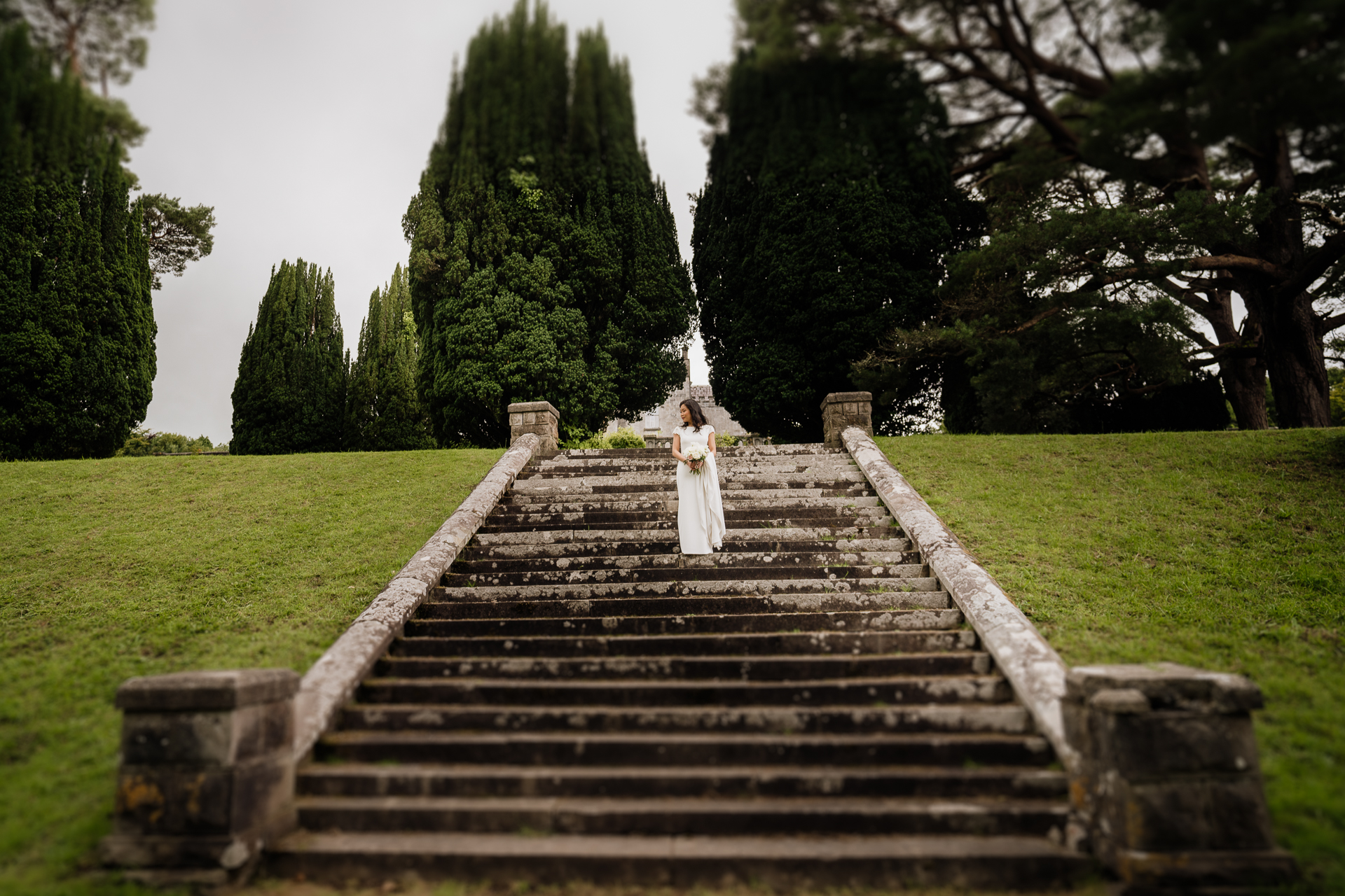 Bride standing alone on the stone steps of Belleek Castle surrounded by tall trees