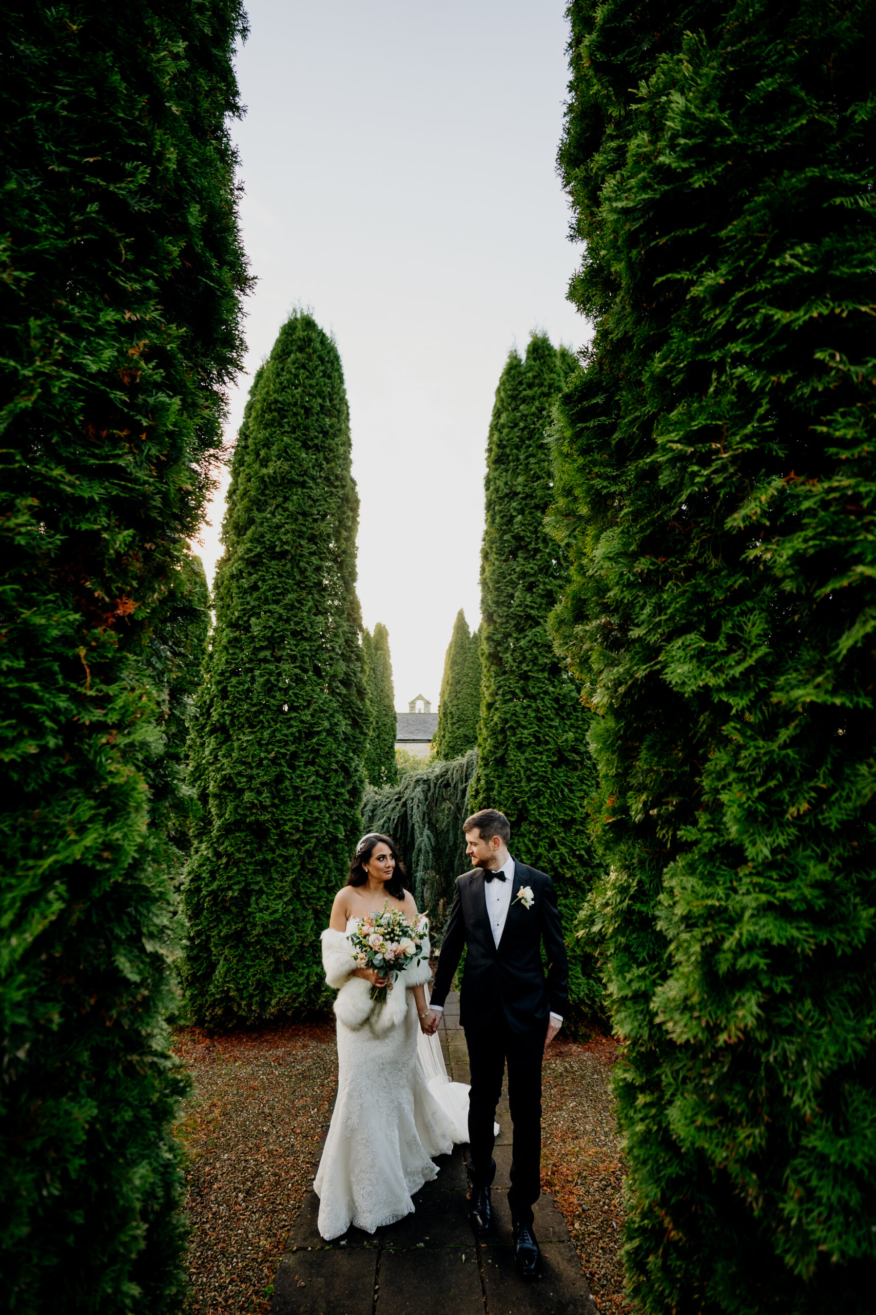 Bride and groom walking hand in hand through a hedge-lined garden path at Cabra Castle