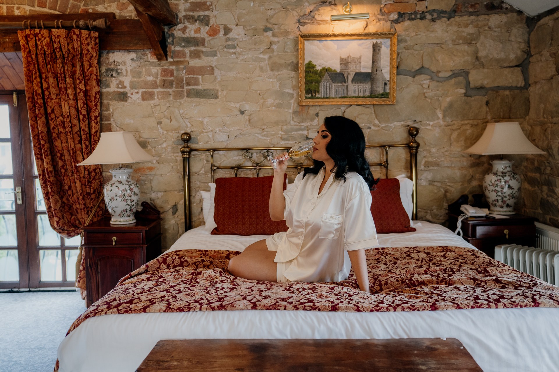 Bride in a silk robe sitting on a bed in a rustic bedroom at Cabra Castle during a winter wedding morning