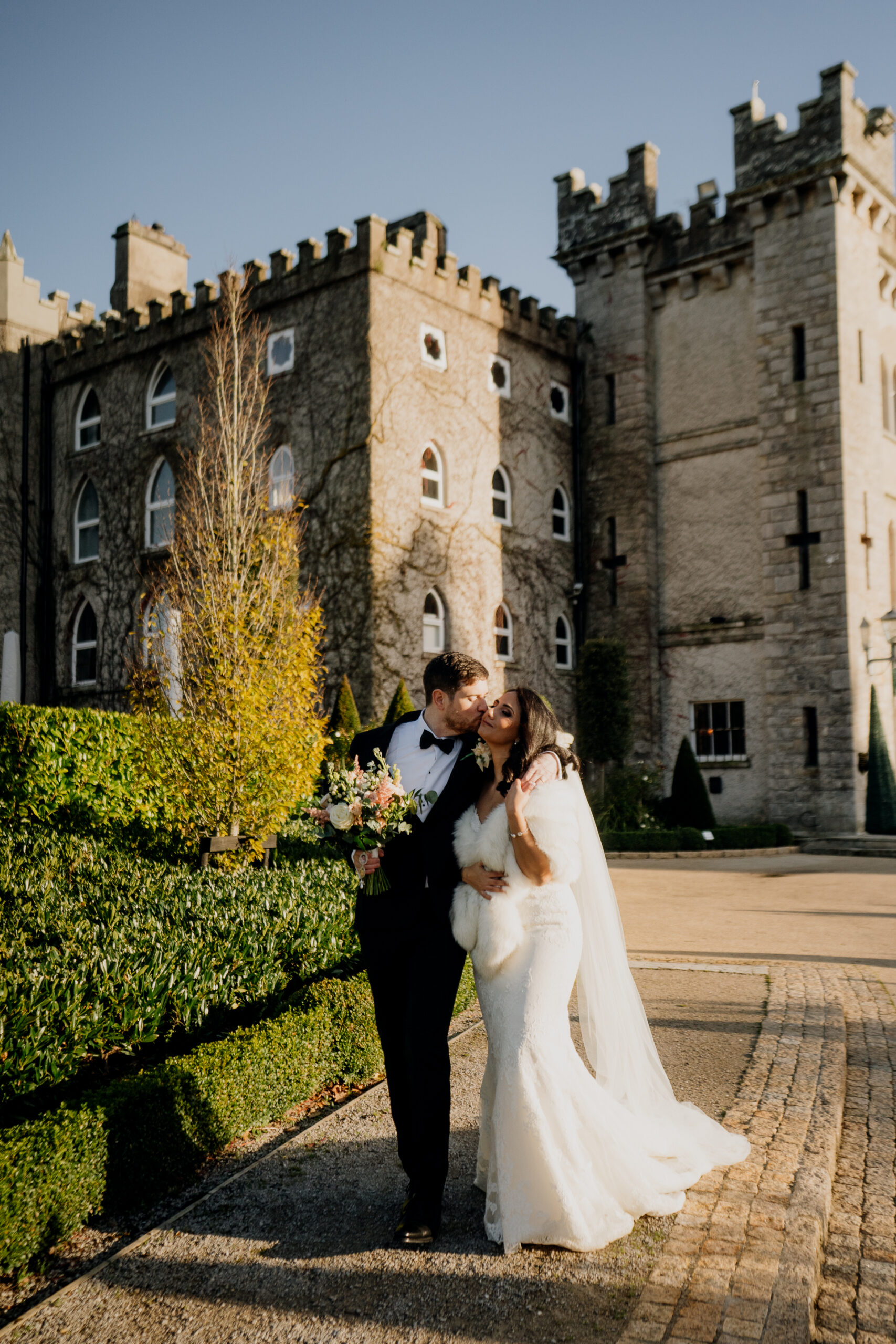 Bride and groom share a candid kiss outside Cabra Castle, walking along the stone path in front of the castle walls.