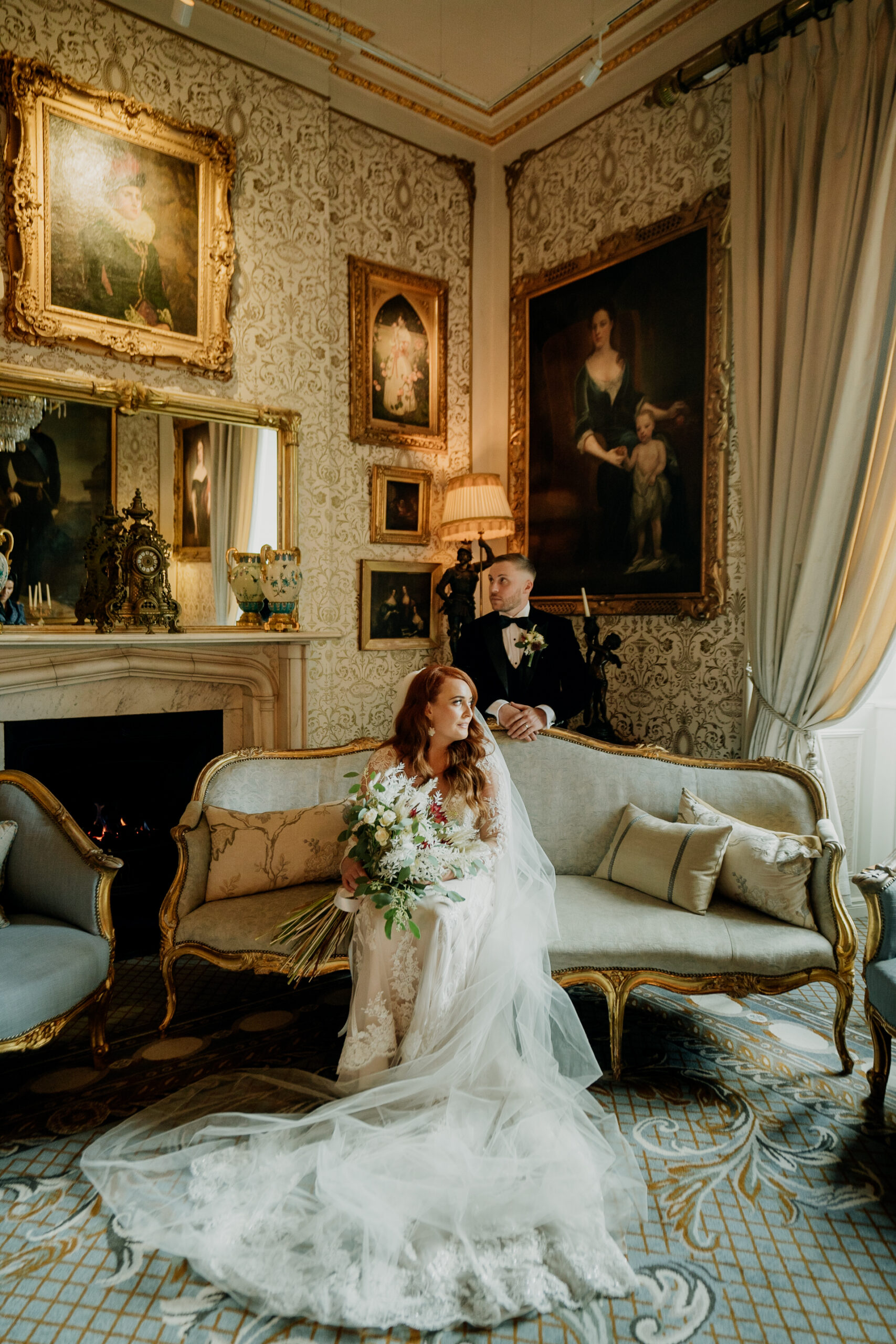 Bride in a wedding gown sitting gracefully on the couch in Cabra Castle’s historic drawing room, surrounded by antique portraits.