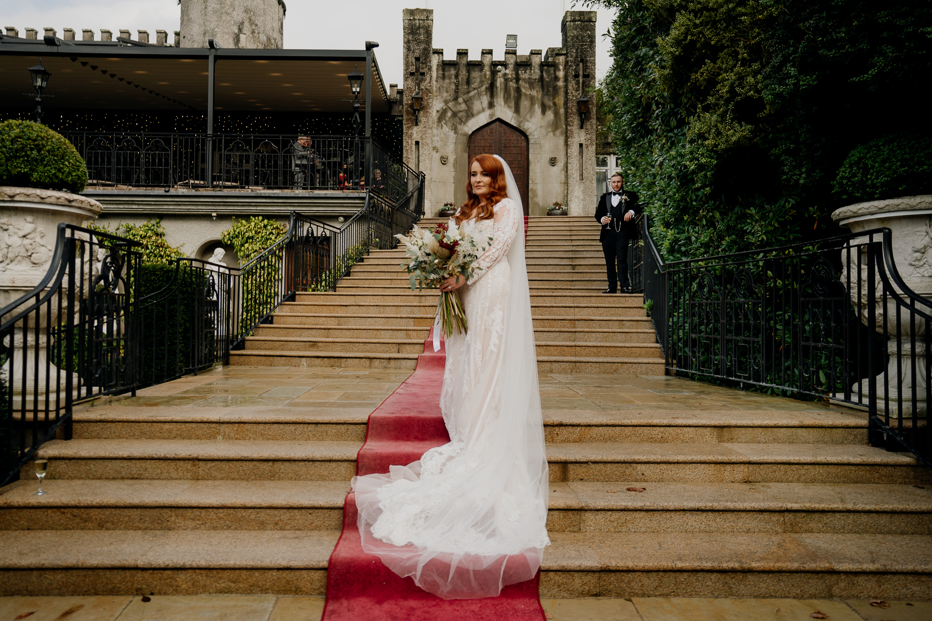 Bride standing alone on the red carpet stairs in front of Cabra Castle, holding her bouquet and looking radiant.