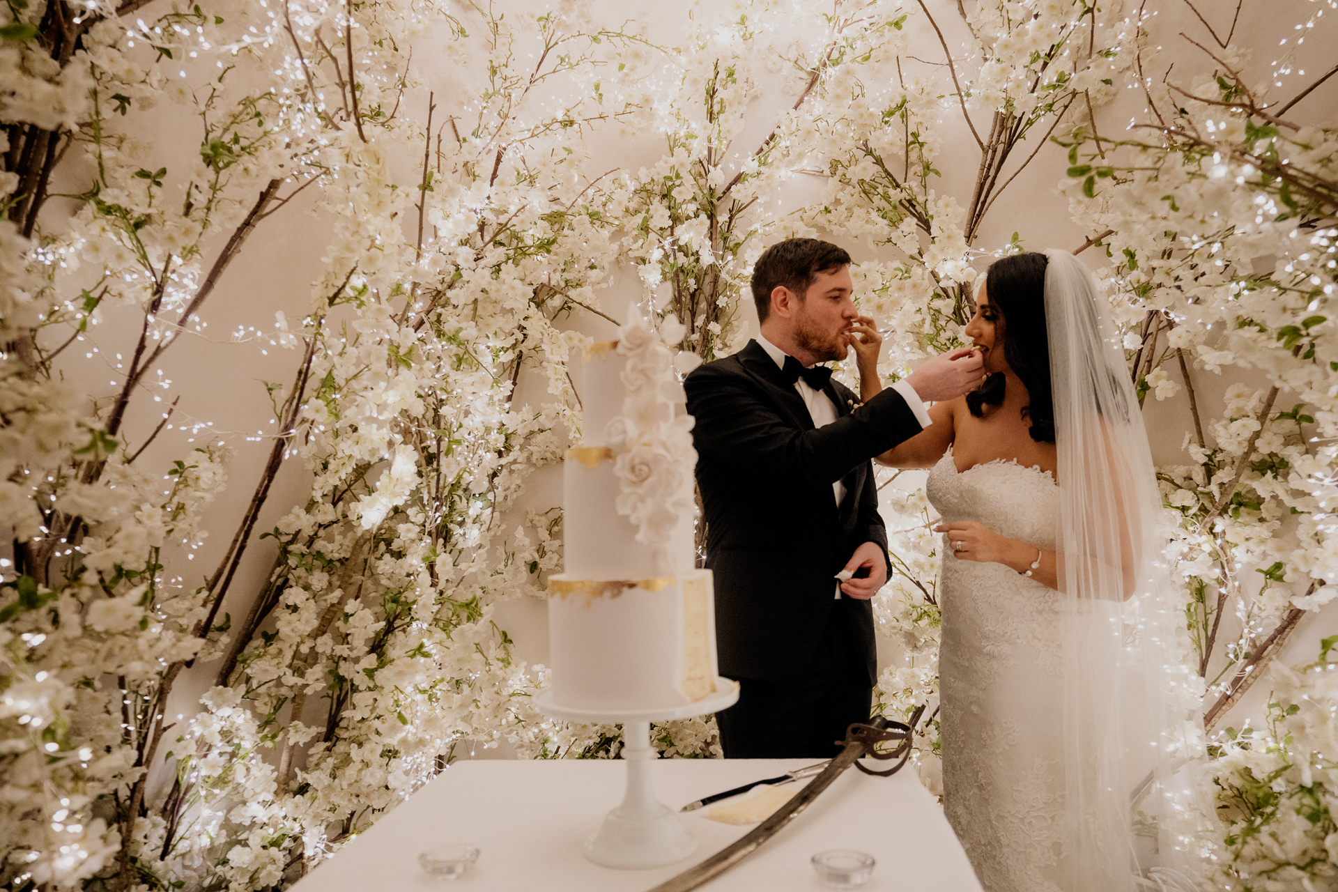 A bride and groom sharing a kiss during their wedding cake cutting under a romantic floral arch at Cabra Castle