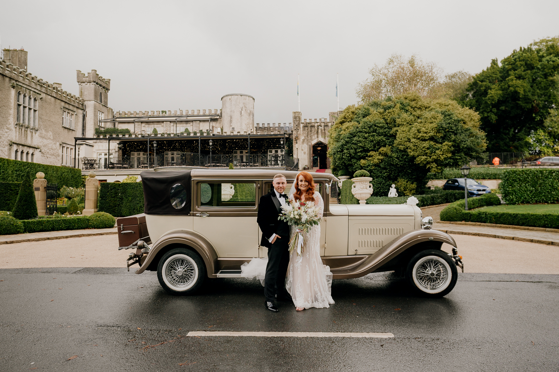 Bride and groom kissing beside a vintage wedding car at Cabra Castle driveway