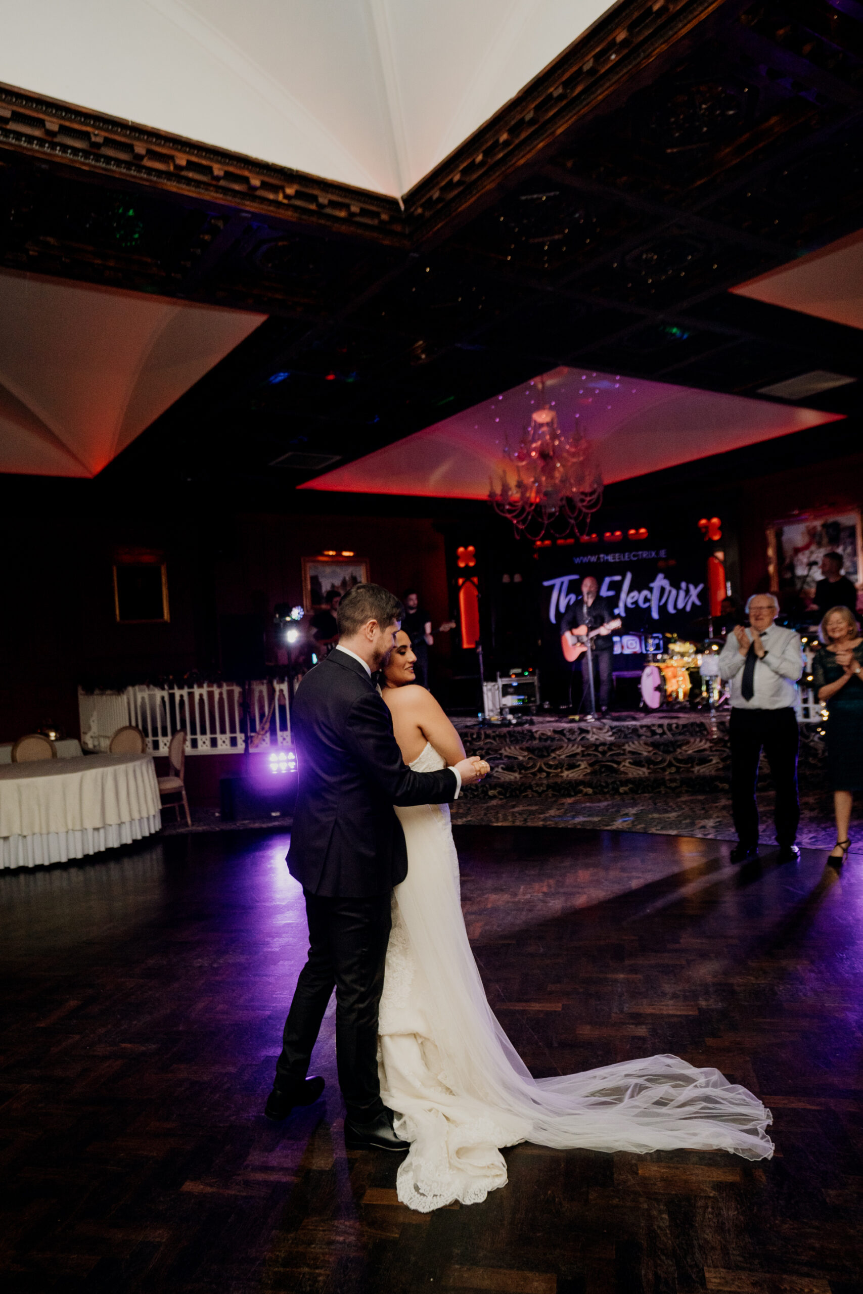 Bride and groom share their first dance on the dark-lit dance floor at Cabra Castle, with dramatic uplighting and guests in the background.