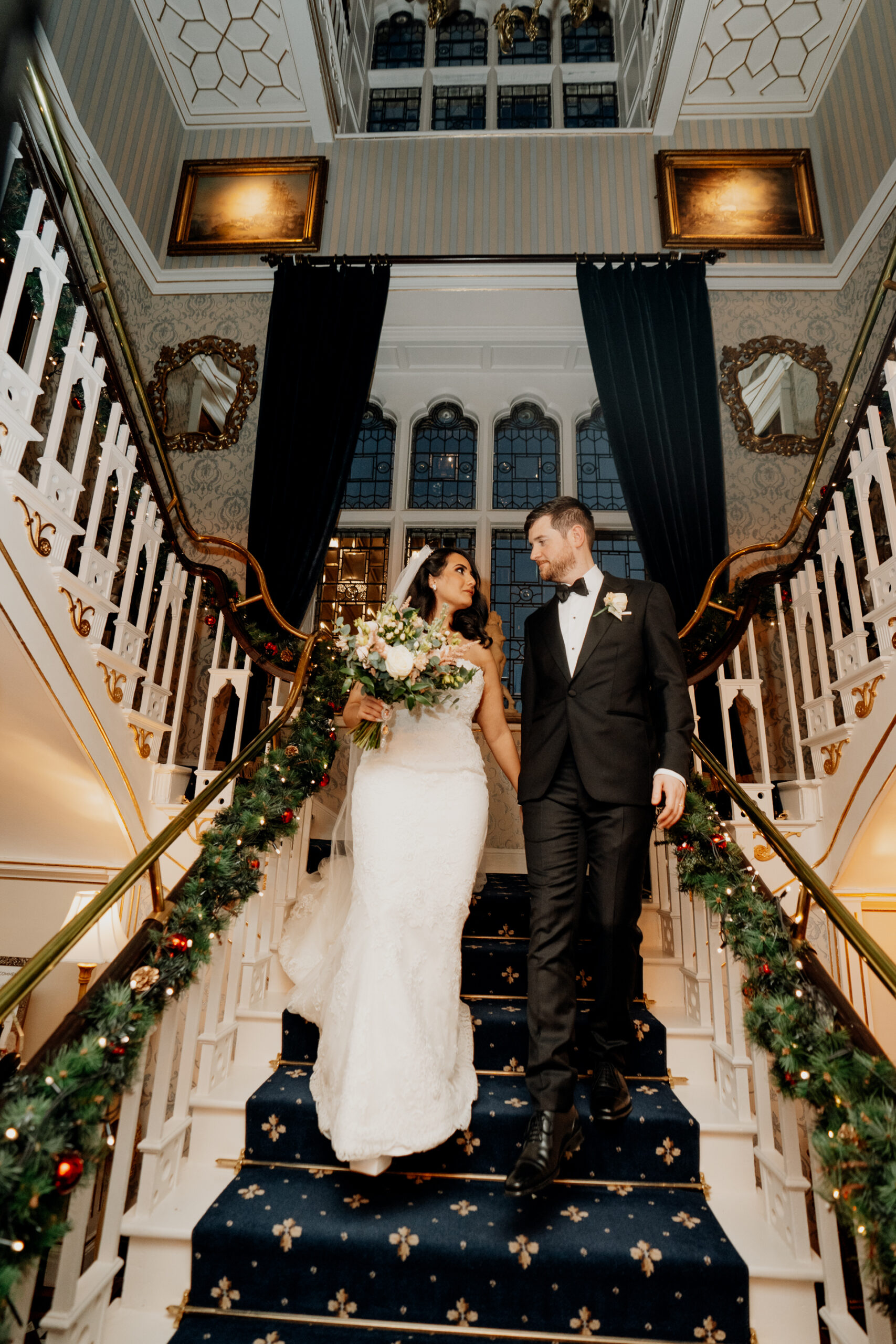 A bride and groom standing together on a grand staircase decorated with greenery and candles at Cabra Castle