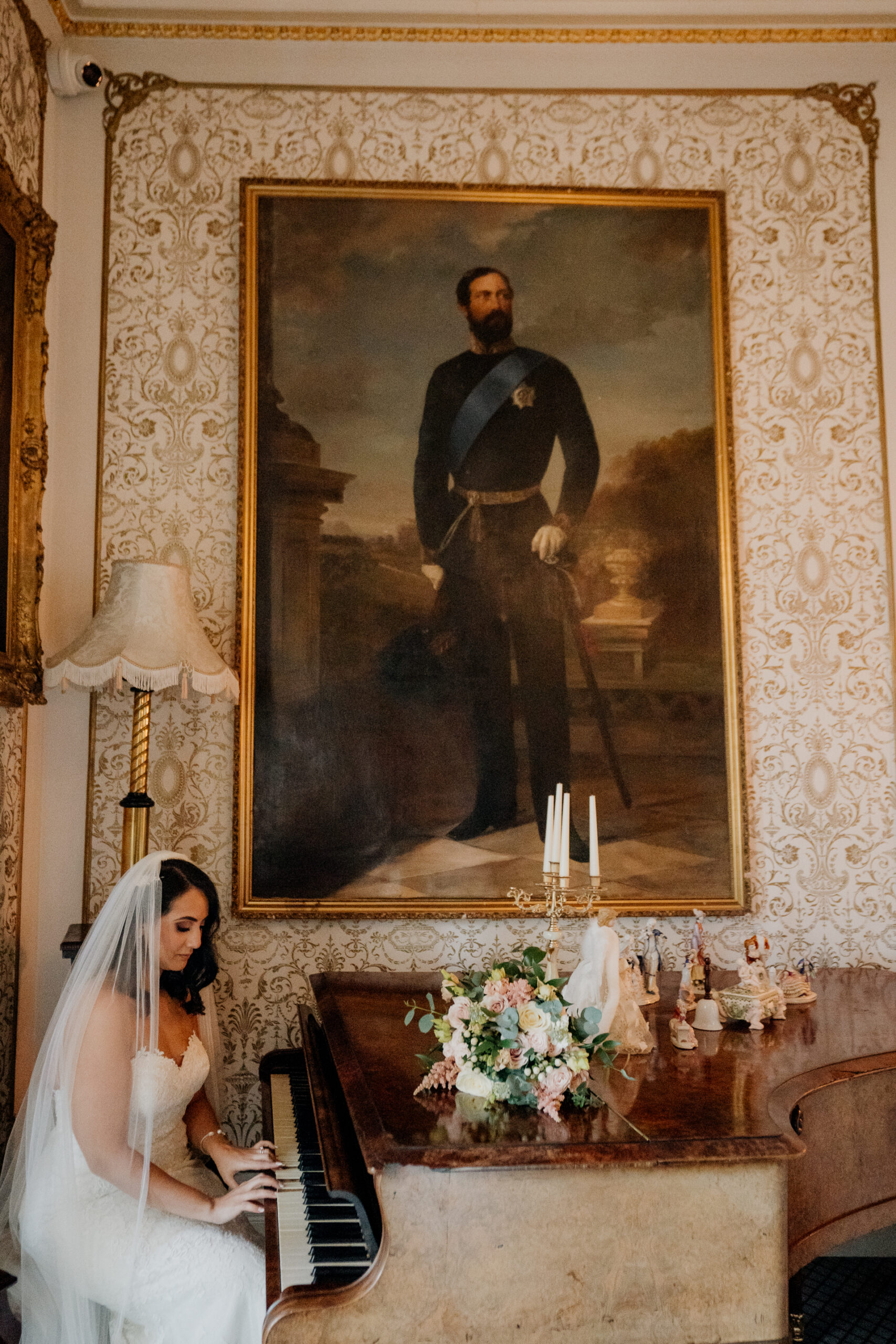 A bride in a white dress seated at a vintage piano under a grand portrait at Cabra Castle