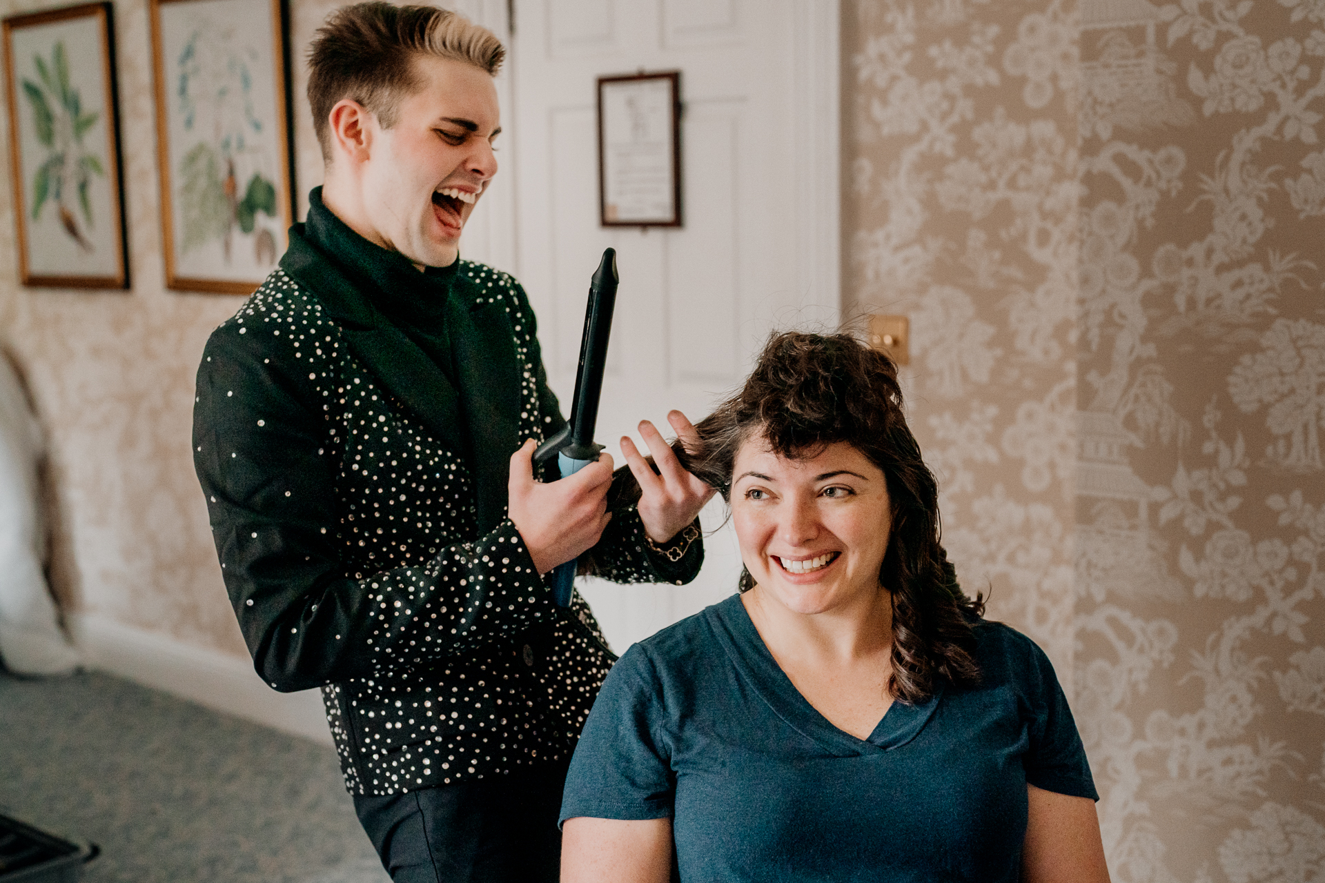 Bridesmaid styling bride’s hair while she sips champagne and laughs