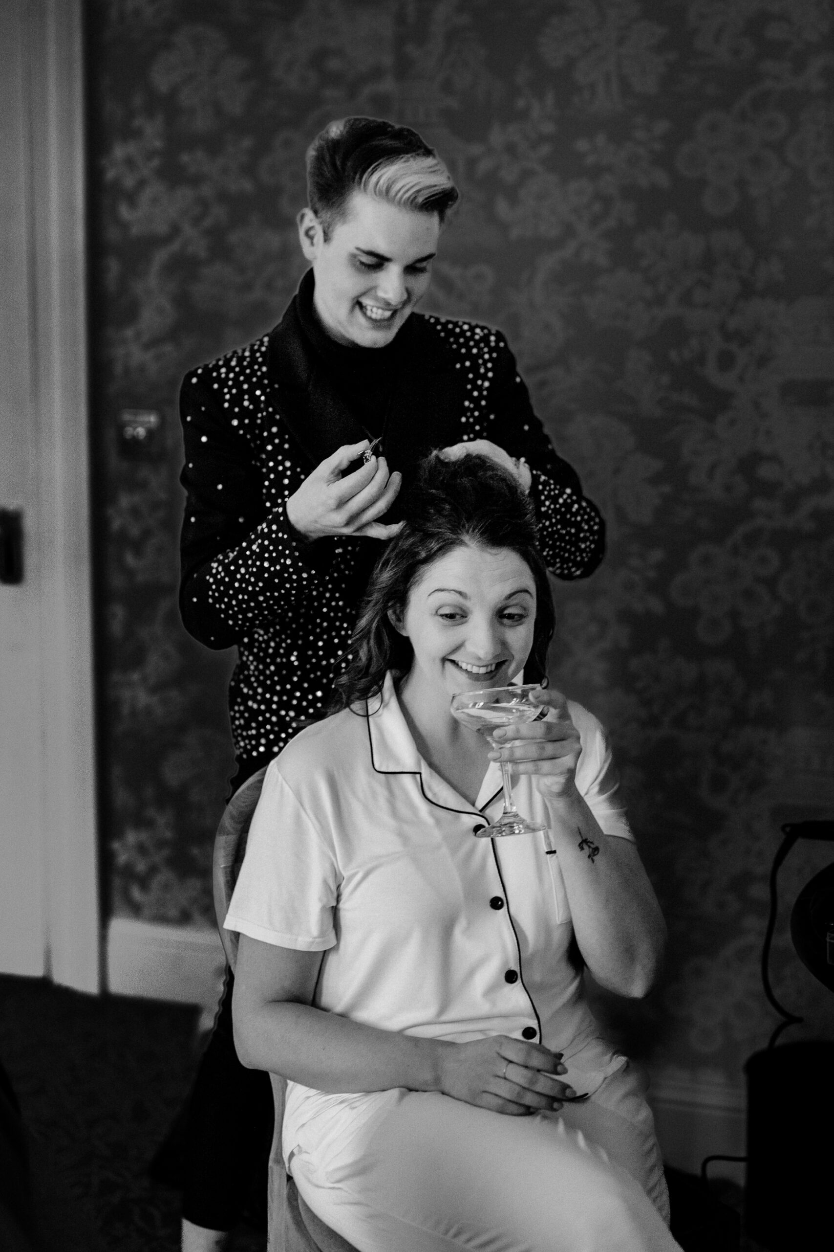 Bridesmaid styling bride’s hair while she sips champagne and laughs
