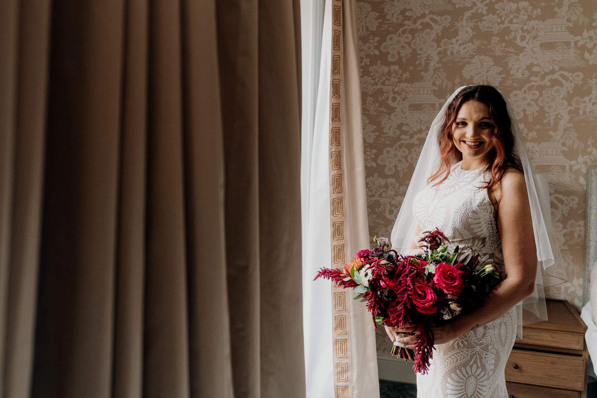 Bride smiling with red bouquet in soft window light before Dublin wedding ceremony