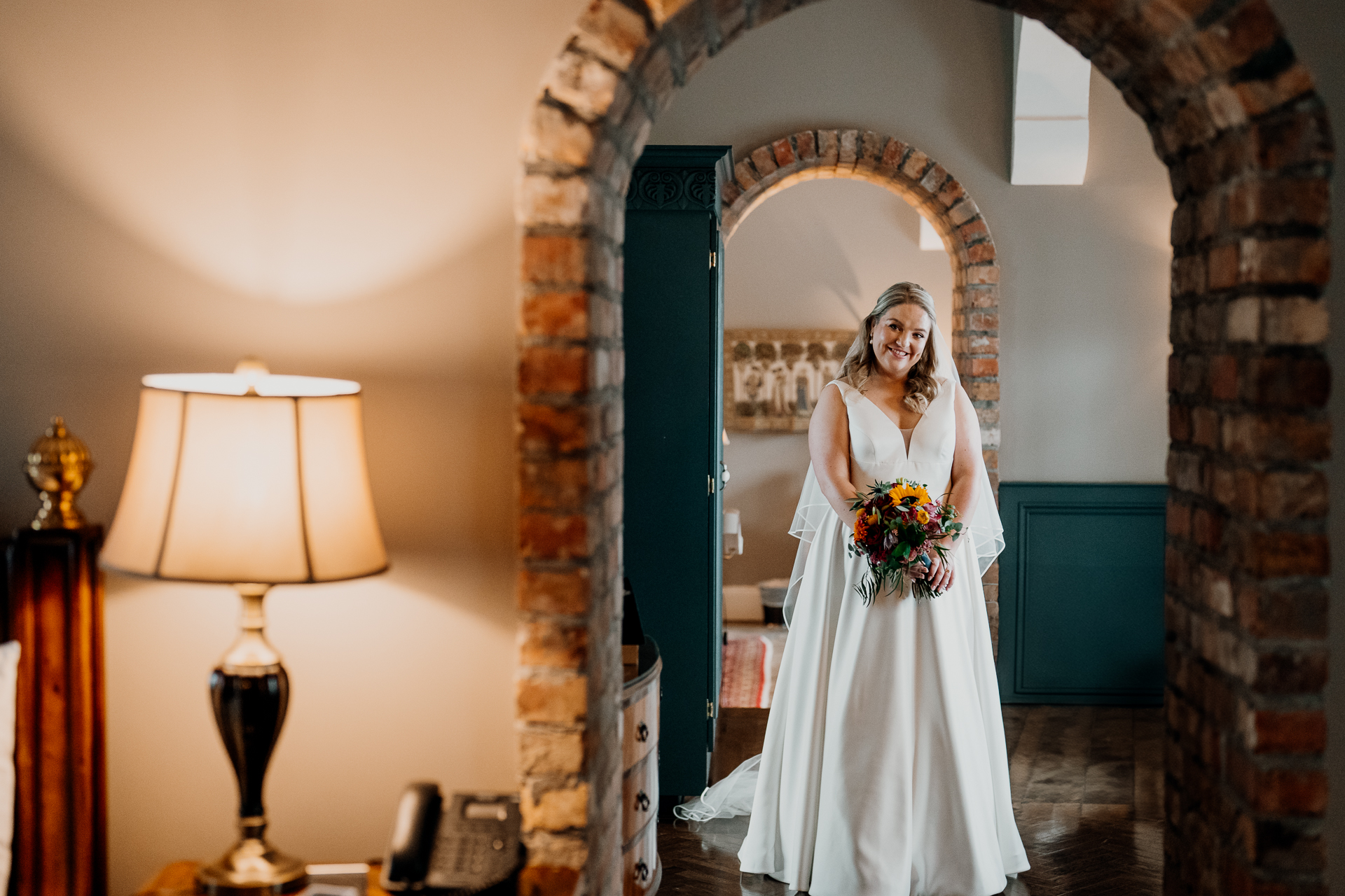 Bride standing under a brick archway in the bridal suite of Markree Castle, holding a bouquet and smiling before the wedding ceremony.